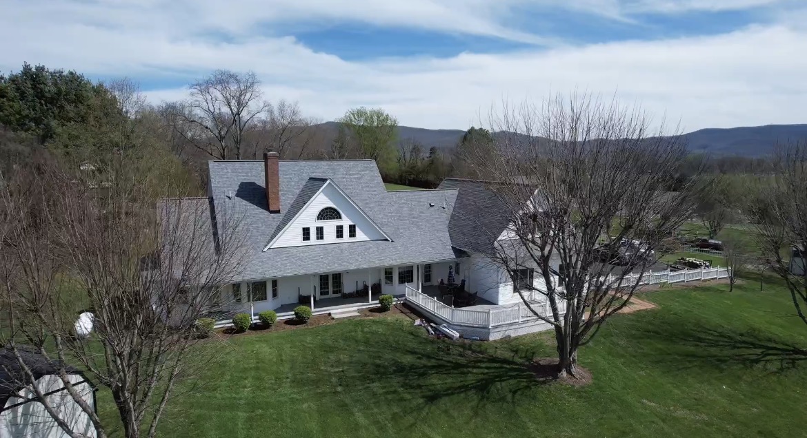 Aerial drone view of white farmhouse-style home with gray shingle roof and mountain backdrop