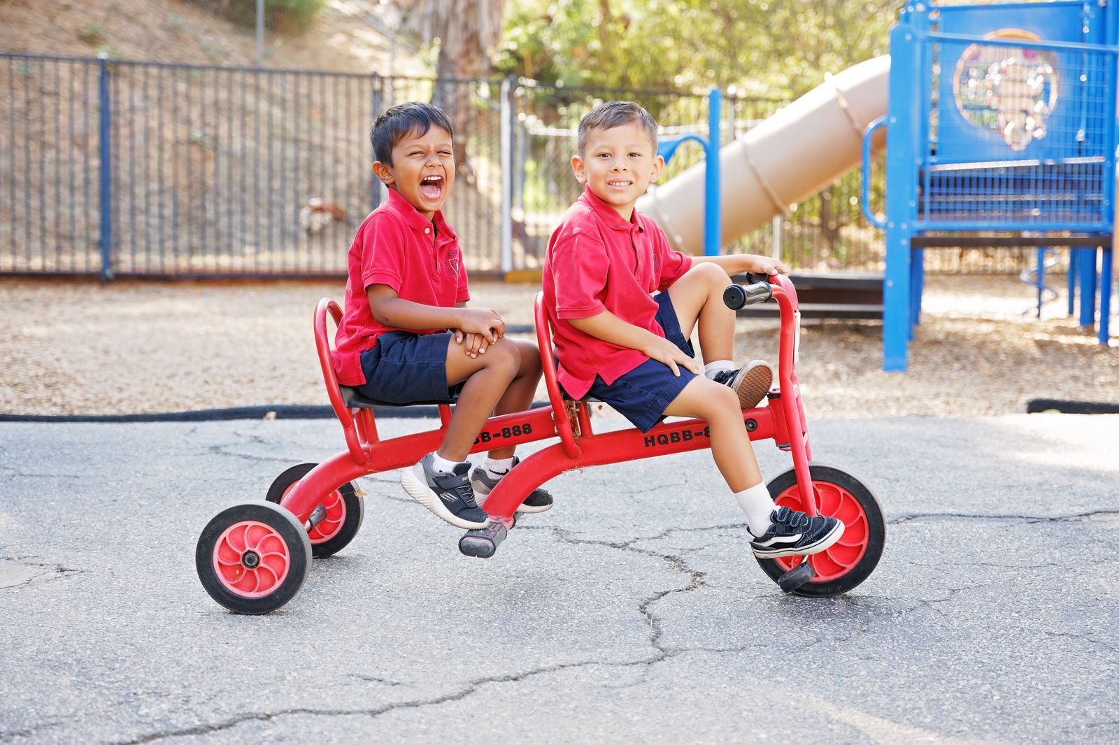 Two young Valley Prep students ride a tandem trike across the playground, laughing