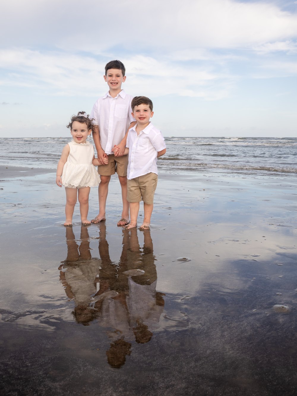 Three children holding hands on the wet sand with their reflections