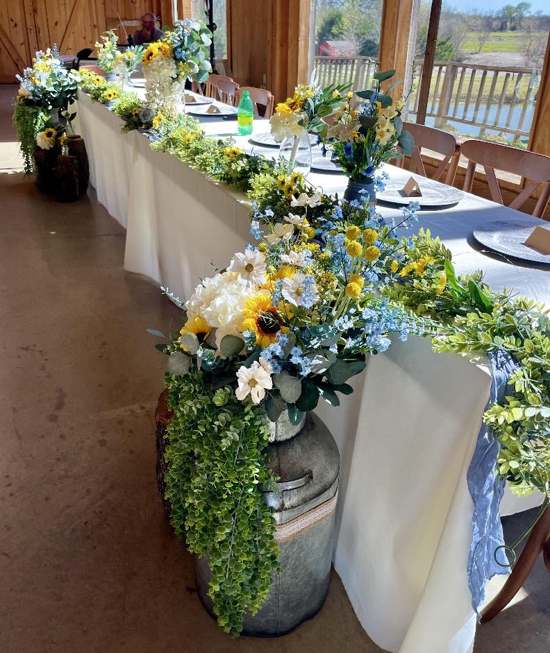 Head table close-up with sunflowers, blue delphinium, cascading greenery, galvanized bucket, and dusty blue runner at DFW wedding