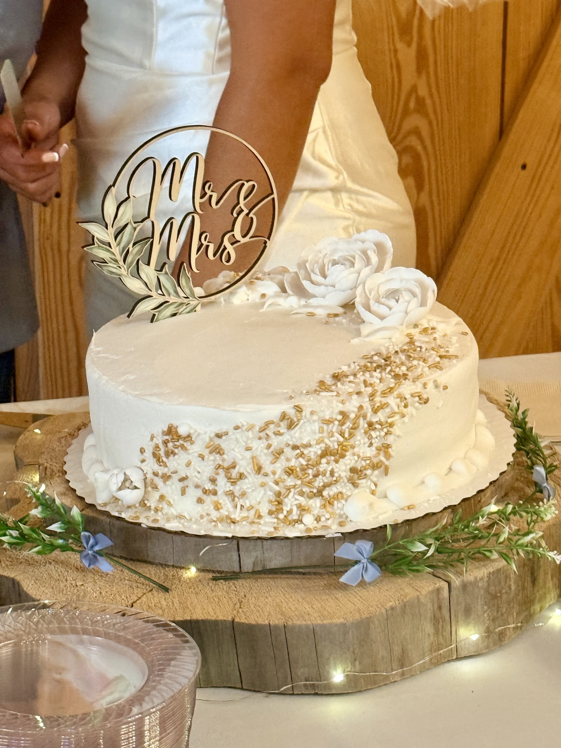 Wedding cake with Mr and Mrs topper, gold sprinkles, white roses, and greenery on rustic wood slab at Trey and Bri wedding in DFW