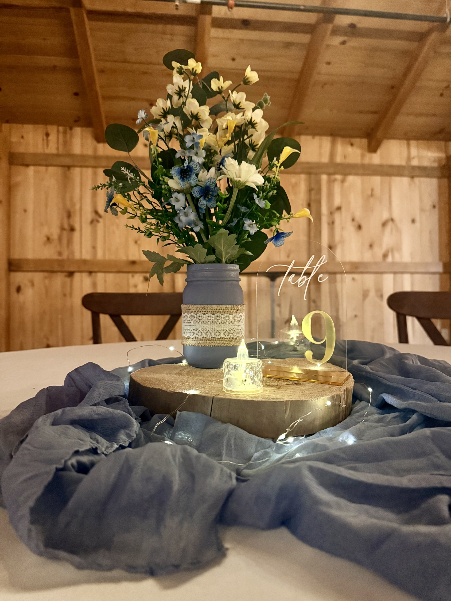 Guest table setup with floral centerpiece on wood slab, dusty blue runner, acrylic table number, and fairy lights at DFW barn wedding