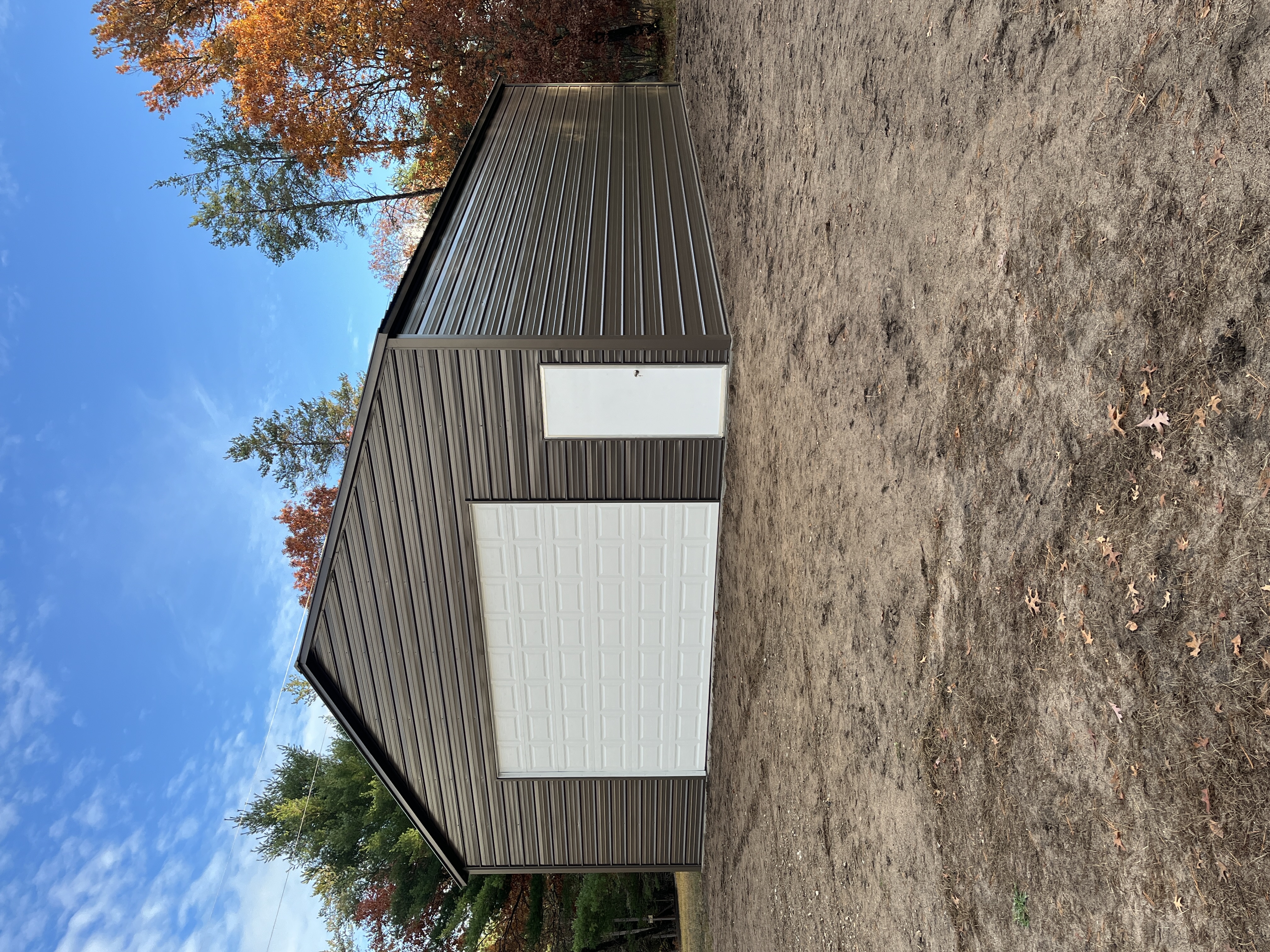 Dark bronze steel building with white garage door and entry door among autumn trees