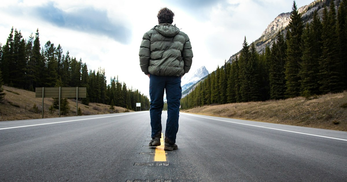 A man stands on the center line of an open road, facing the mountains ahead