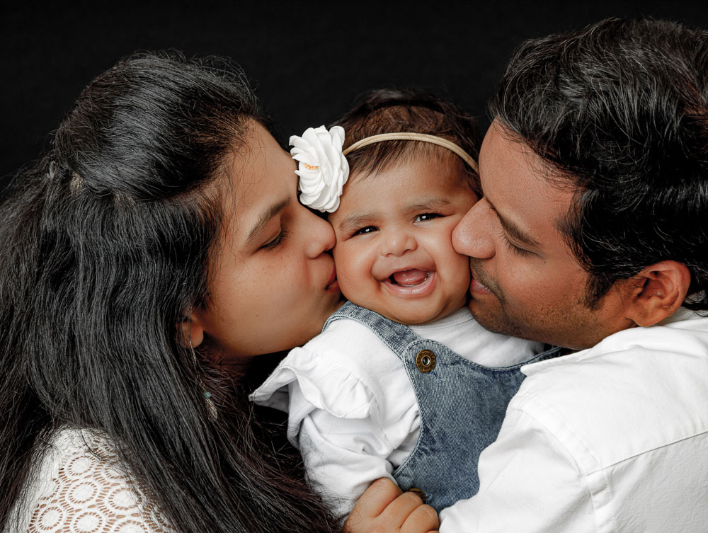 Baby grinning ear-to-ear while both parents lean in to kiss her cheeks — IDTPhoto 3-month sitter session