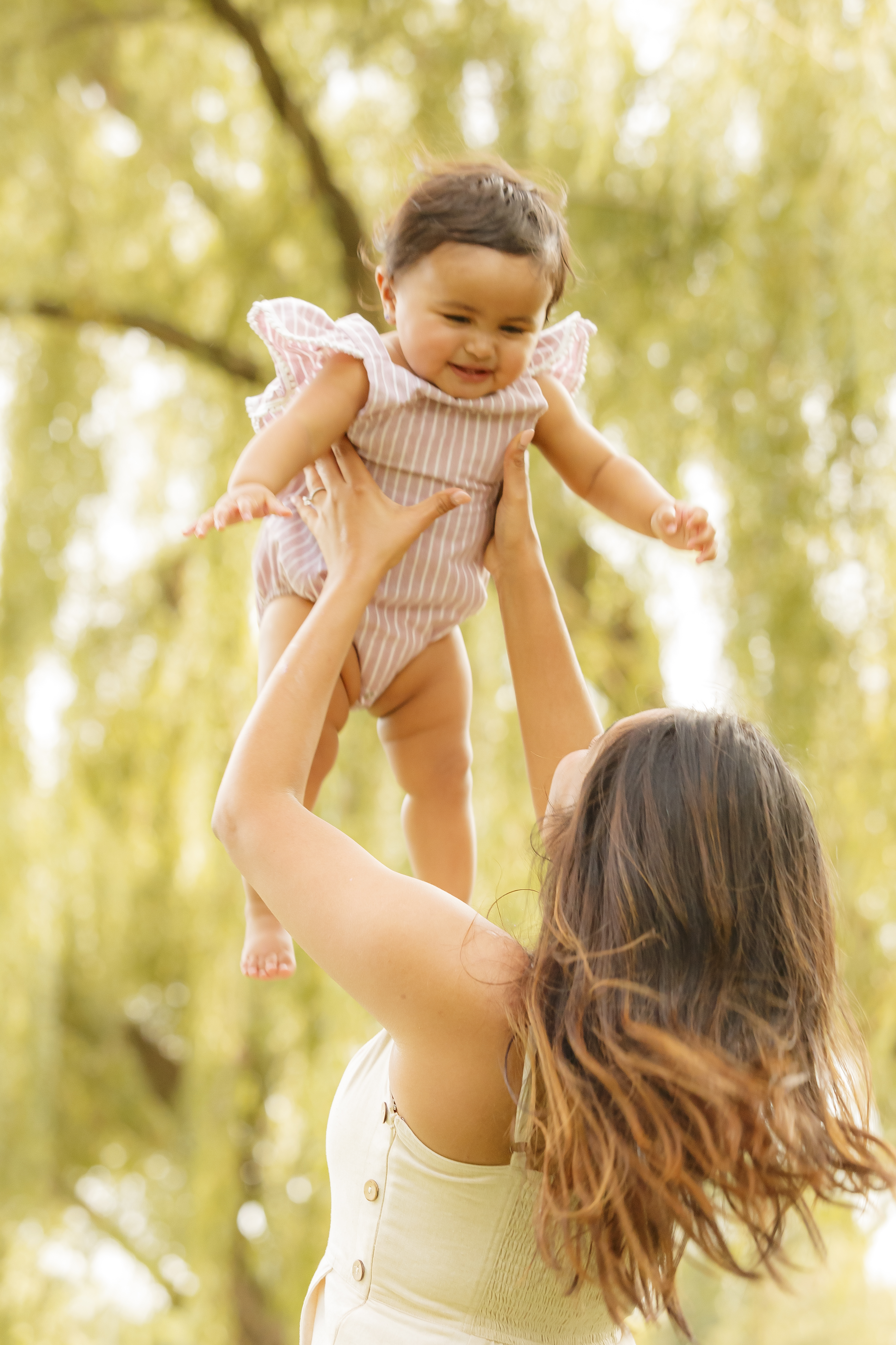 Mom lifting laughing baby high in the air under weeping willow trees outdoors