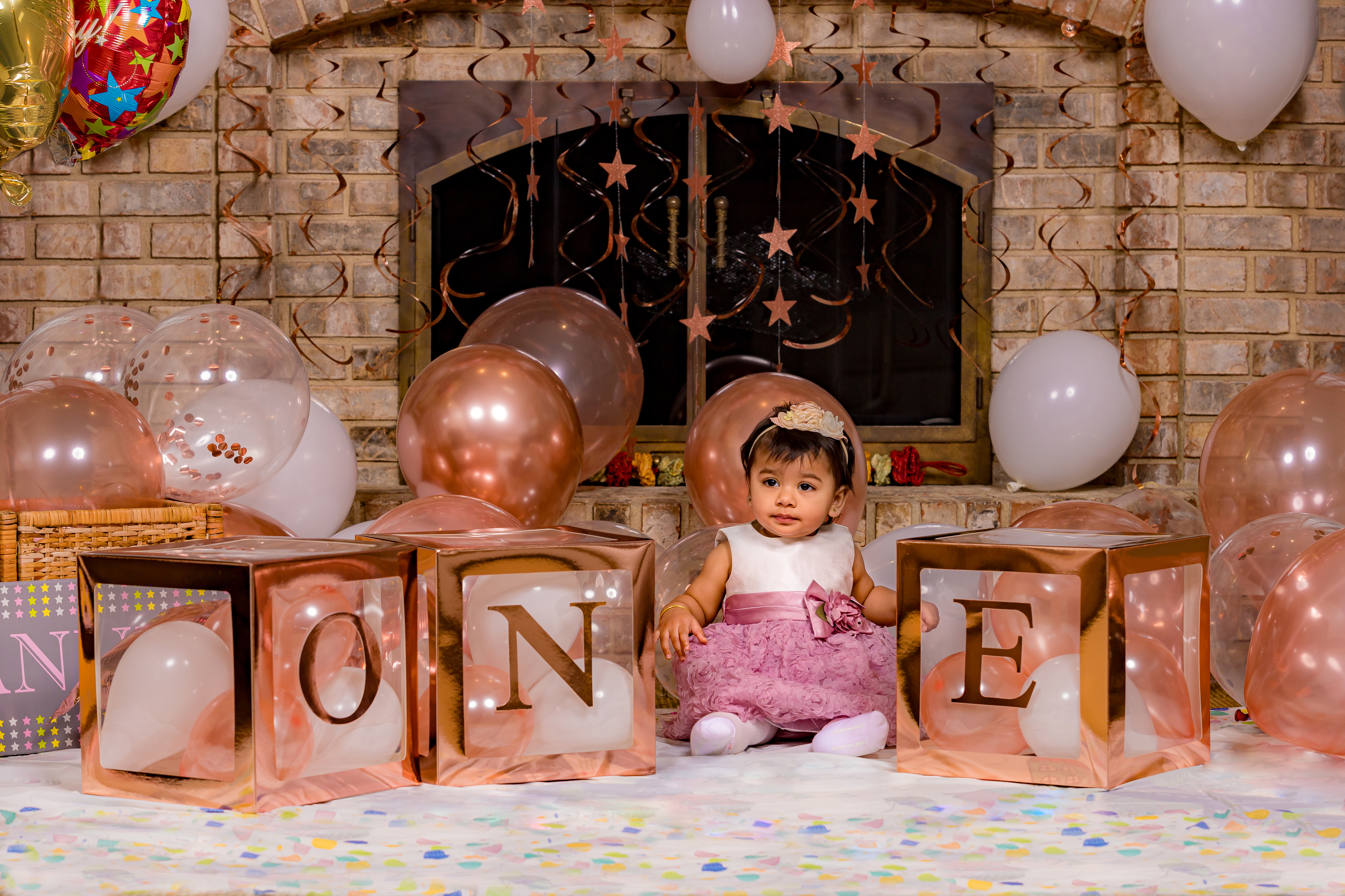 Baby girl sitting between O-N-E letter boxes with rose gold balloons and fireplace backdrop — indoor studio cake smash by IDTPhoto