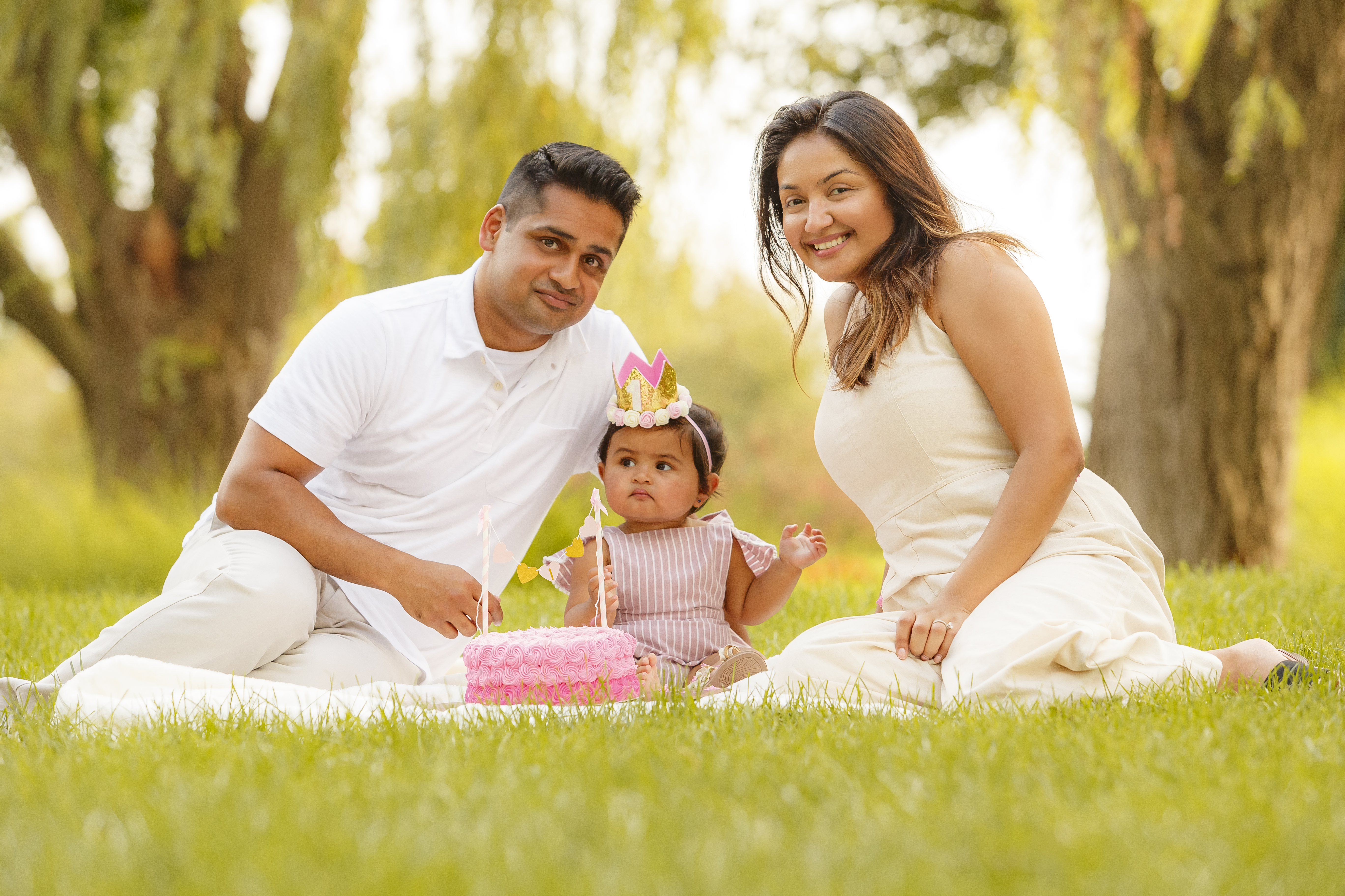 Parents sitting on blanket with baby girl and pink birthday cake during outdoor session
