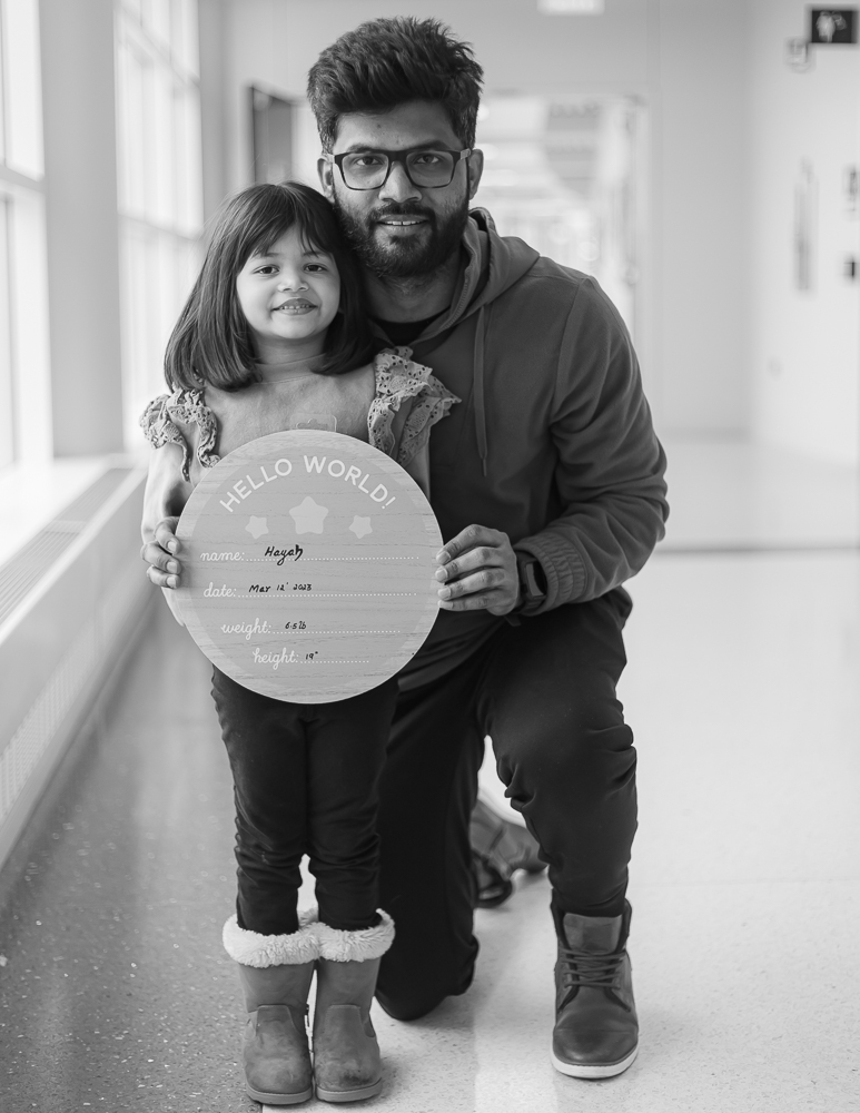 Fresh 48 black & white — dad kneeling in hospital hallway with older sibling holding Hello World birth board