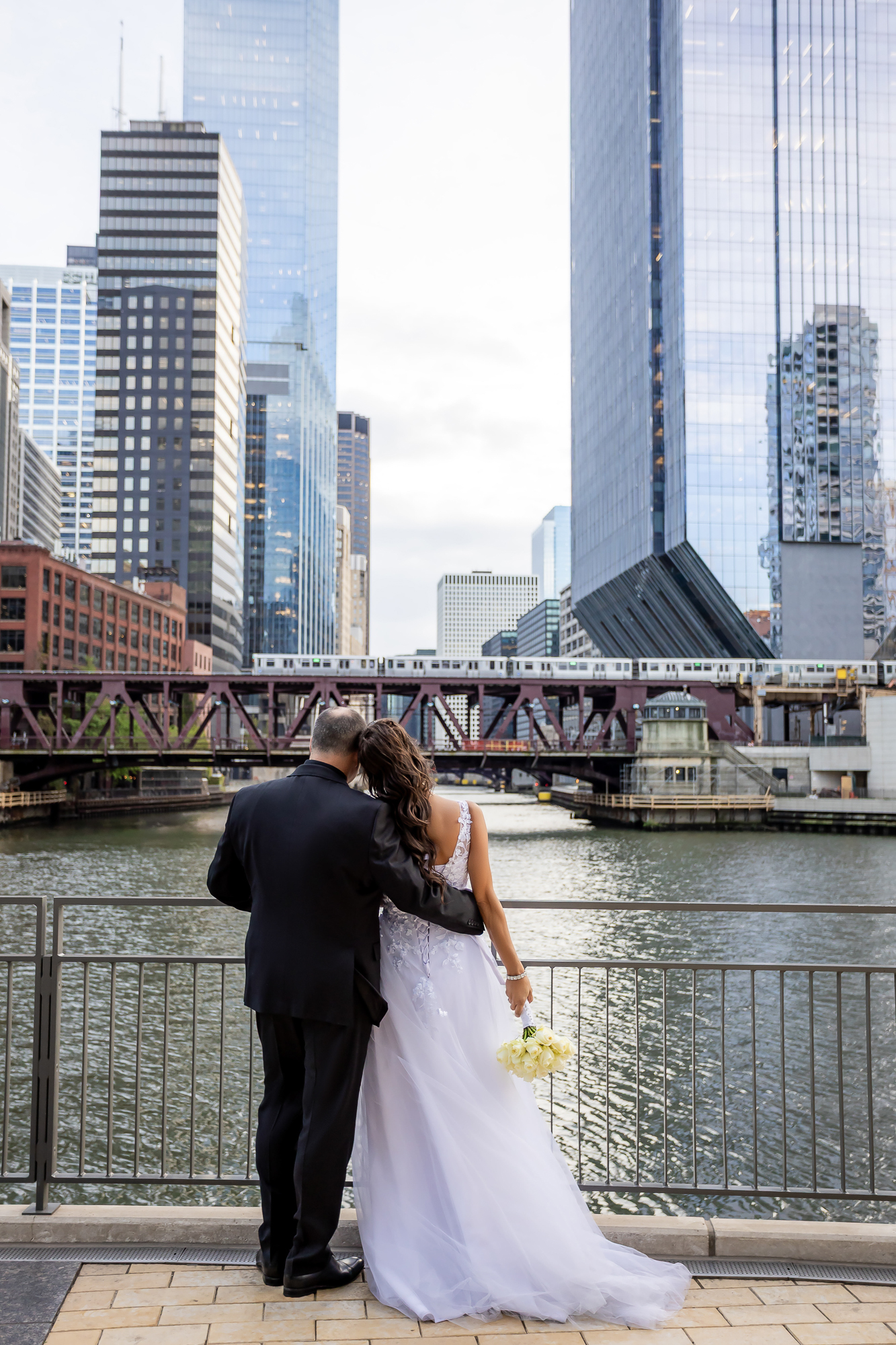 Bride leaning toward groom along Chicago Riverwalk with downtown skyline and L train — IDTPhoto wedding photography