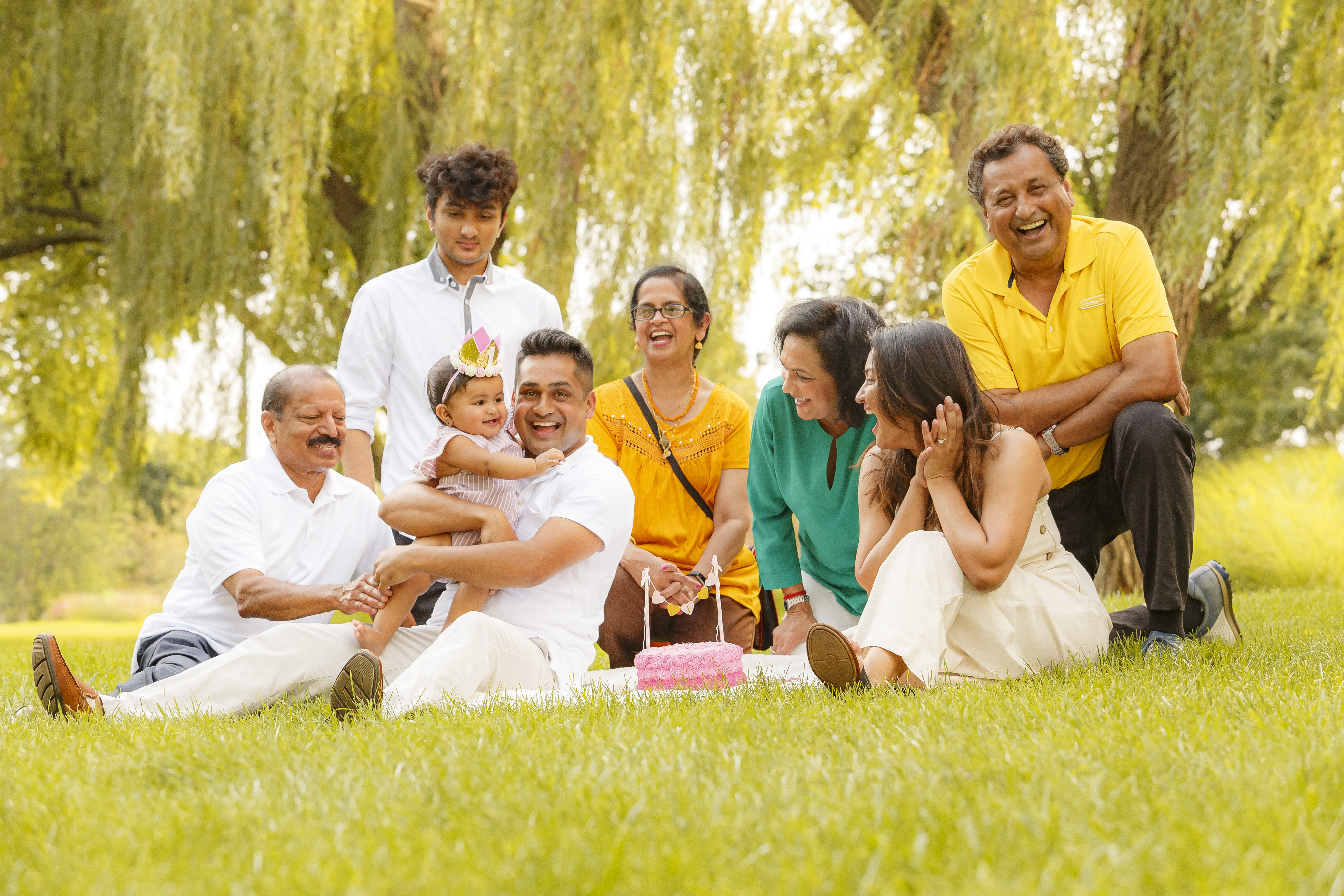 Extended family laughing and celebrating baby's first birthday outdoors under weeping willows