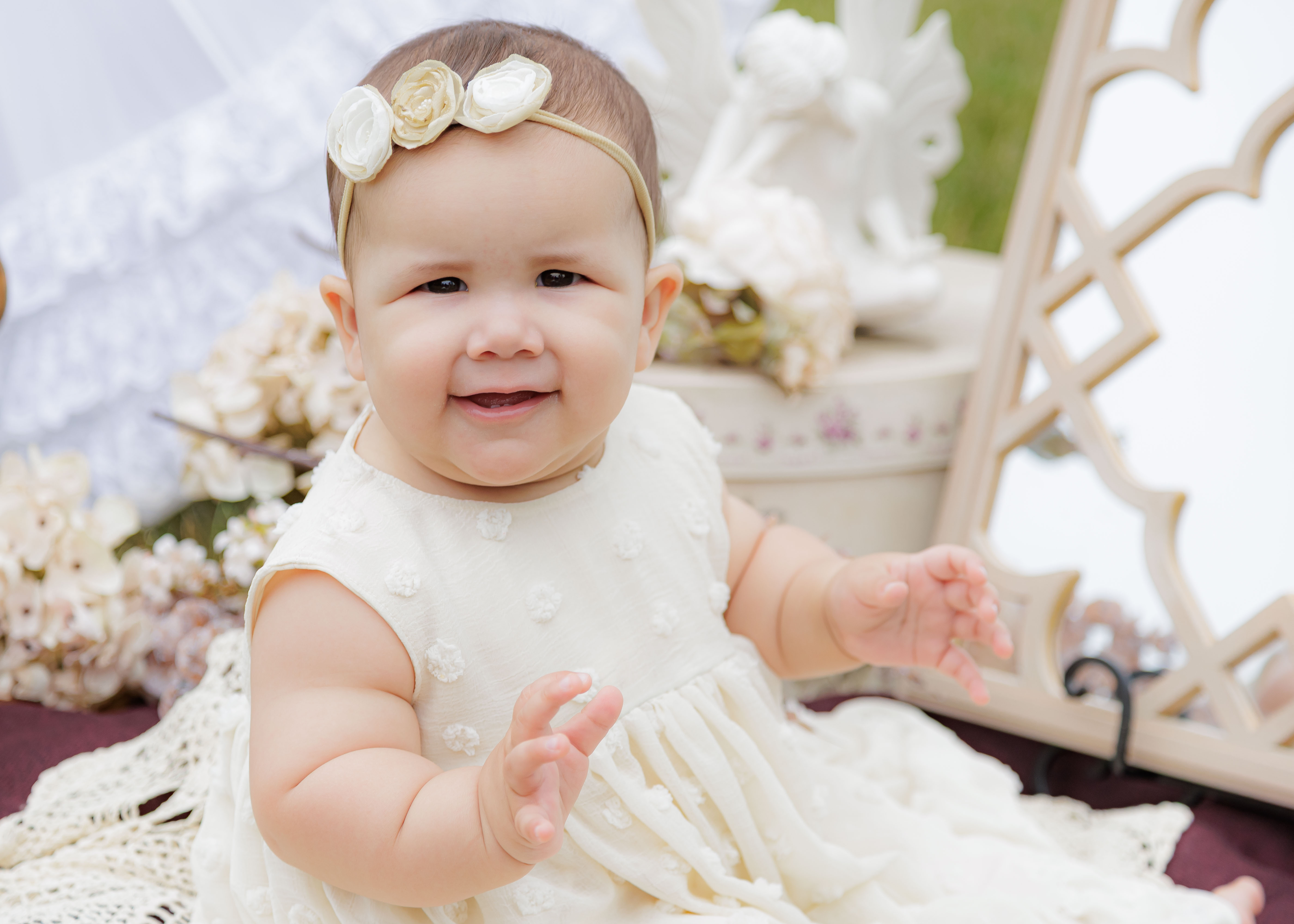 Baby girl in cream dotted dress with triple rose headband smiling and clapping in vintage outdoor garden setup — IDTPhoto sitter session