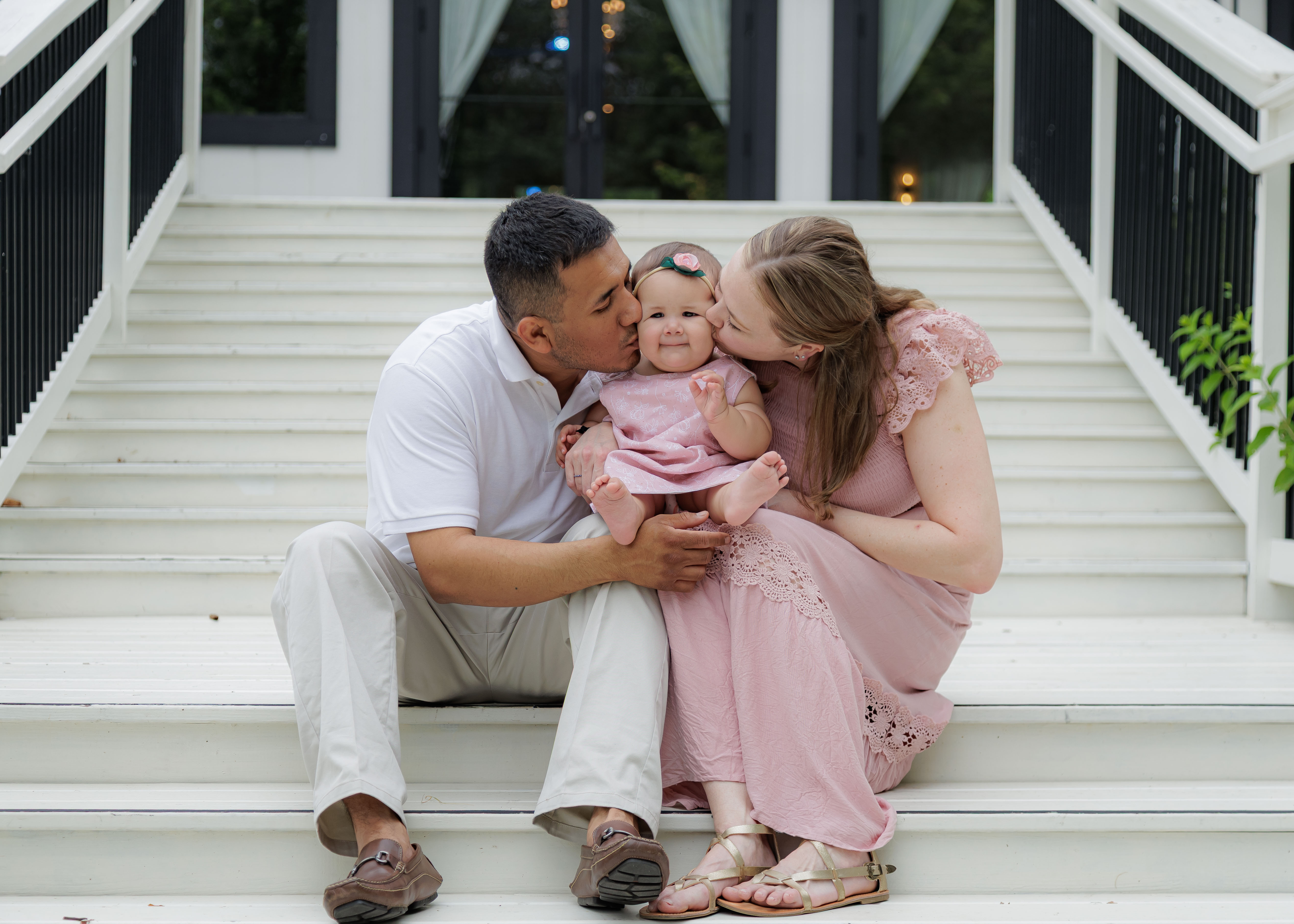 Both parents kissing baby girl's cheeks simultaneously on white outdoor steps — IDTPhoto milestone portrait