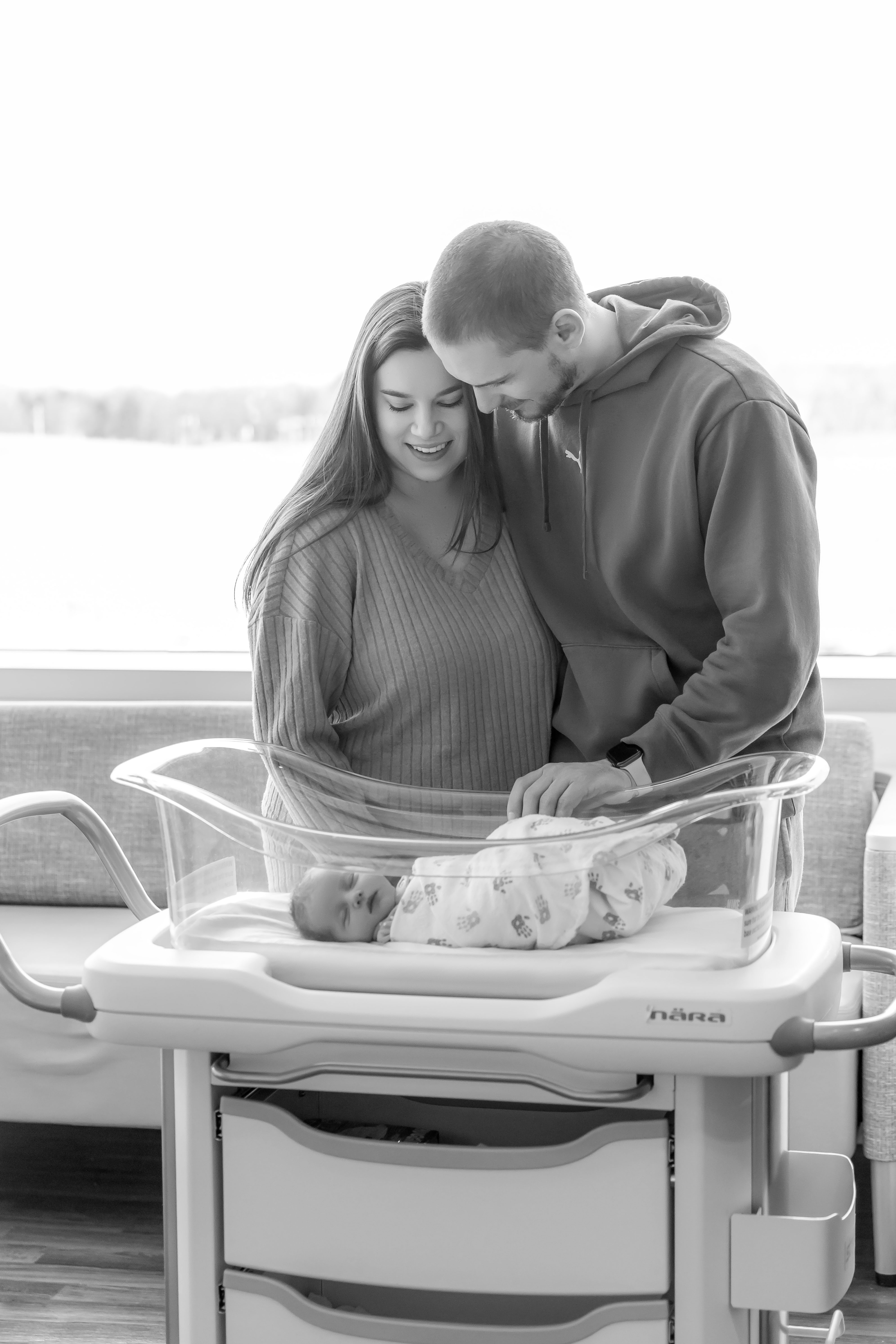Fresh 48 black & white — new parents leaning together over newborn in hospital bassinet