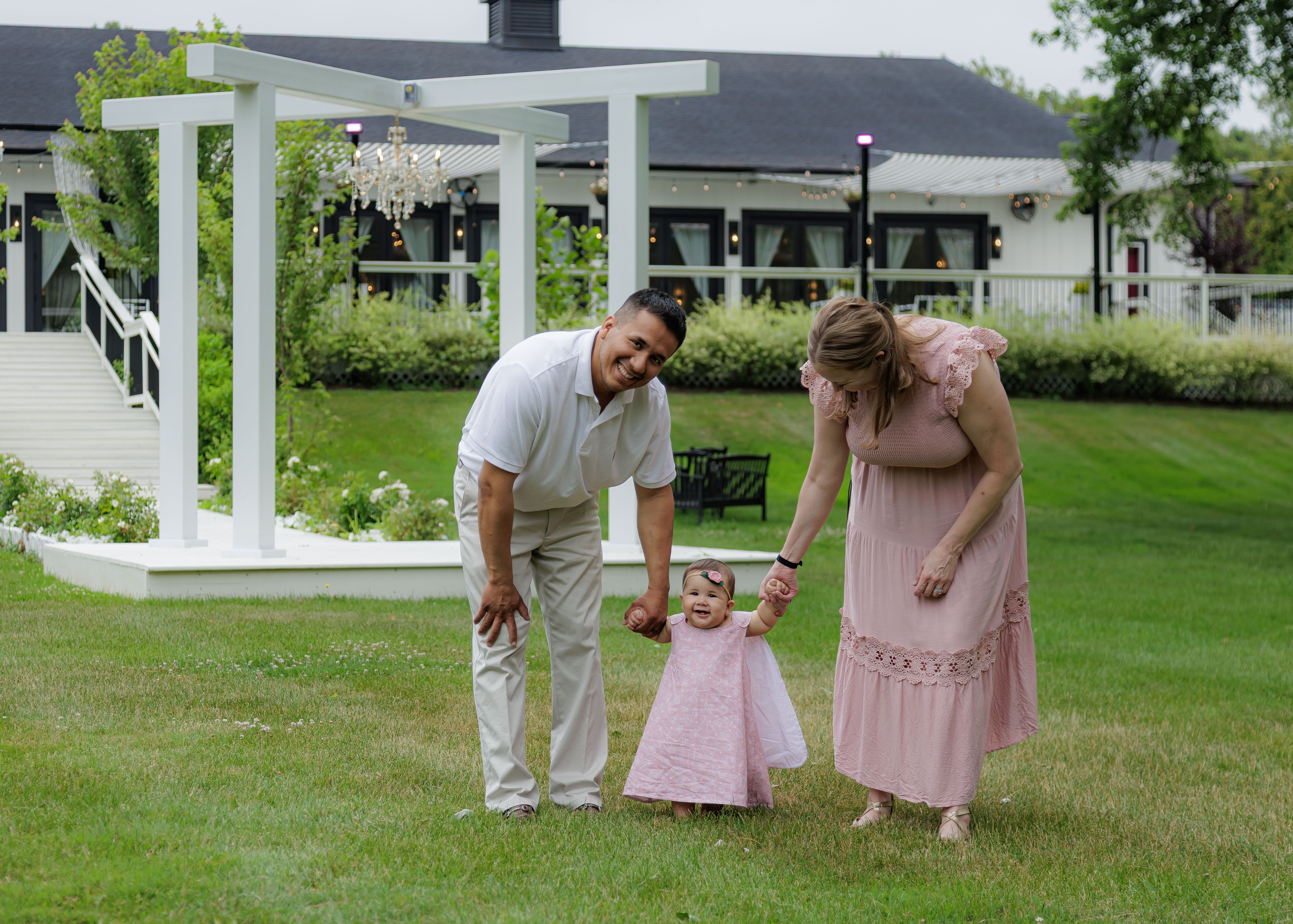Parents helping baby girl take her first steps on venue lawn, both parents smiling with joy — IDTPhoto 9-month milestone session