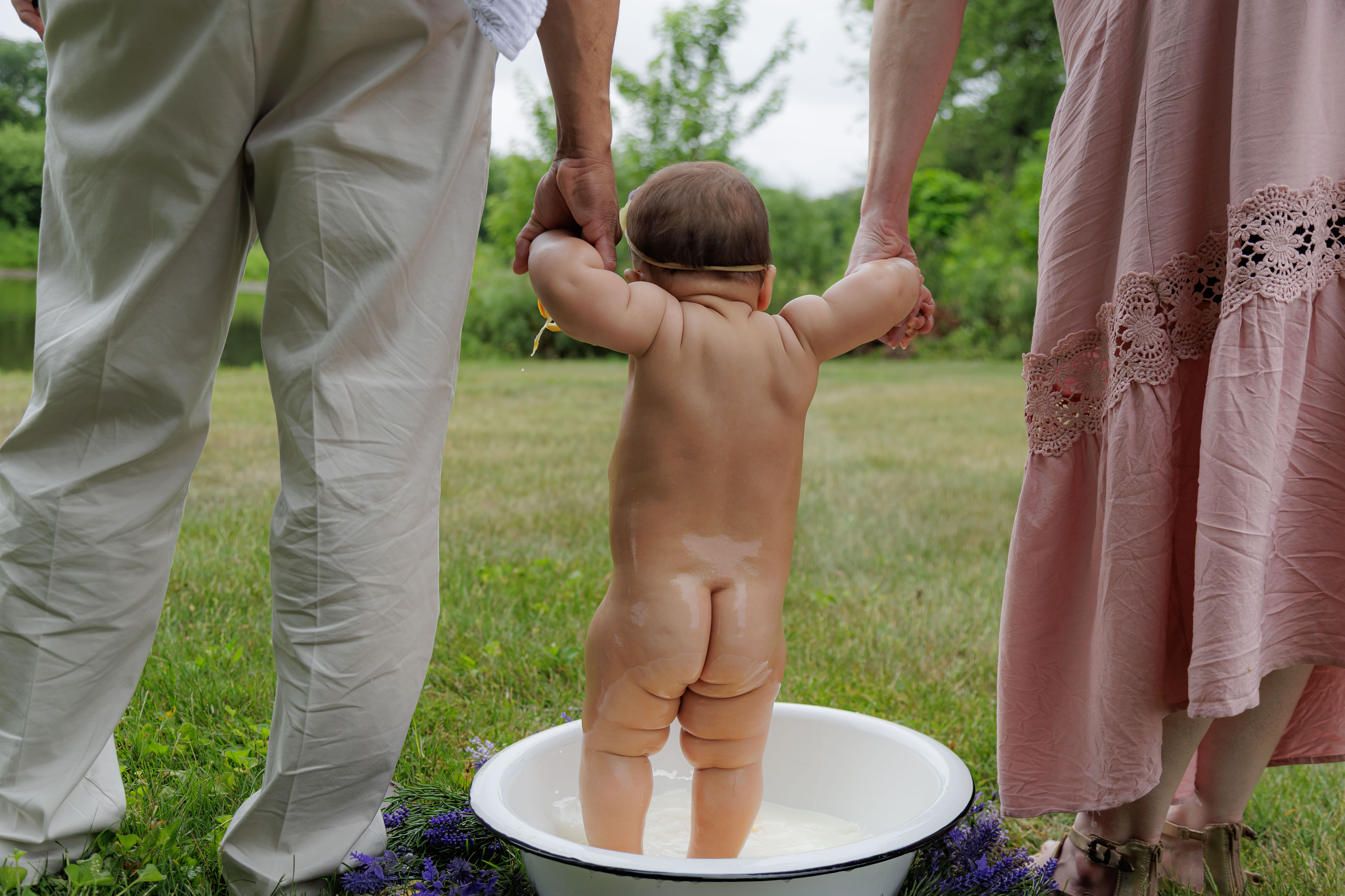 Baby standing in outdoor milk bath tub from behind, parents holding her hands on either side — IDTPhoto milestone session Chicagoland