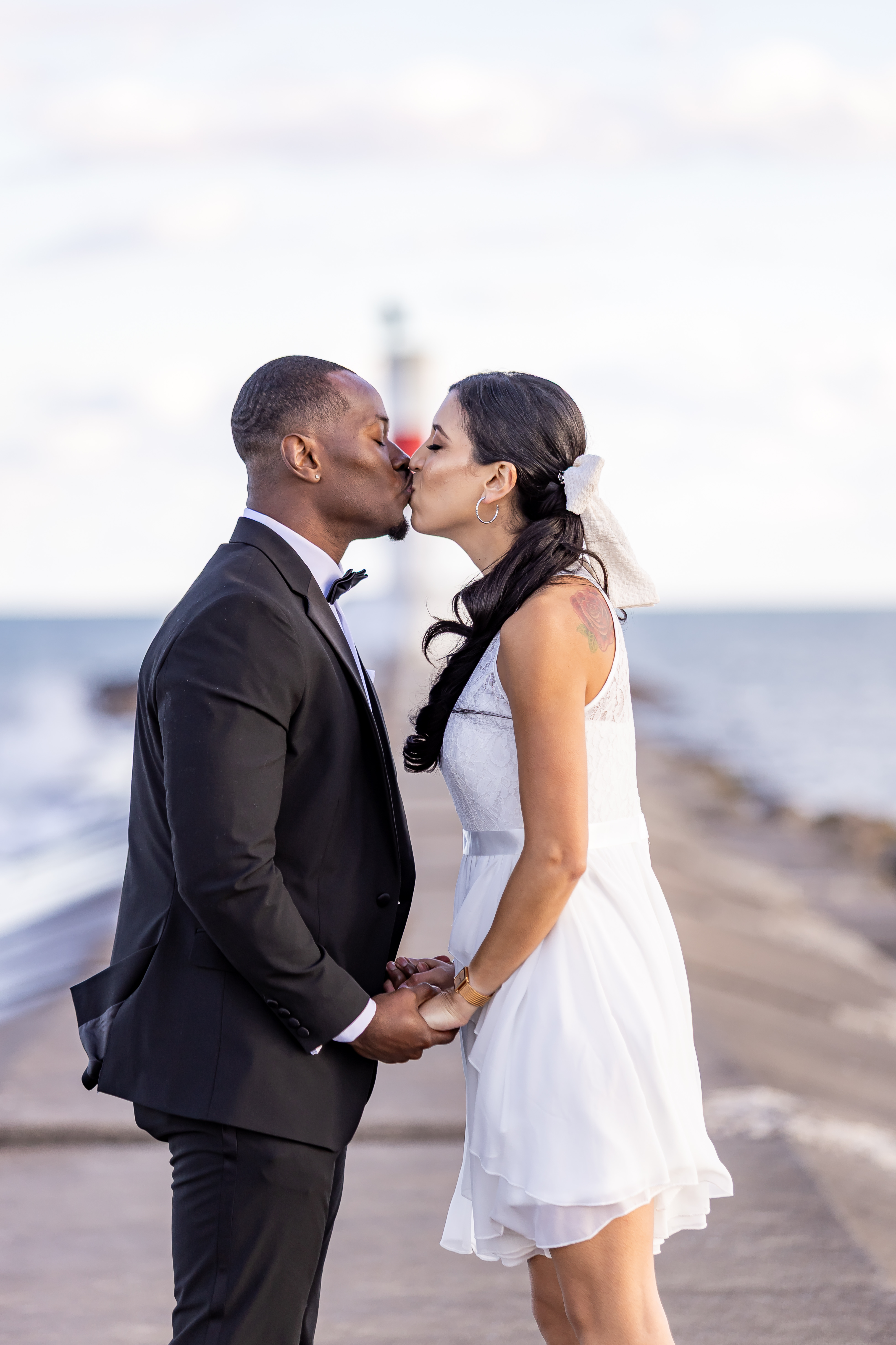 Couple in black wedding attire walking away down beach with footprints in sand and sunflower bouquet — IDTPhoto elopement