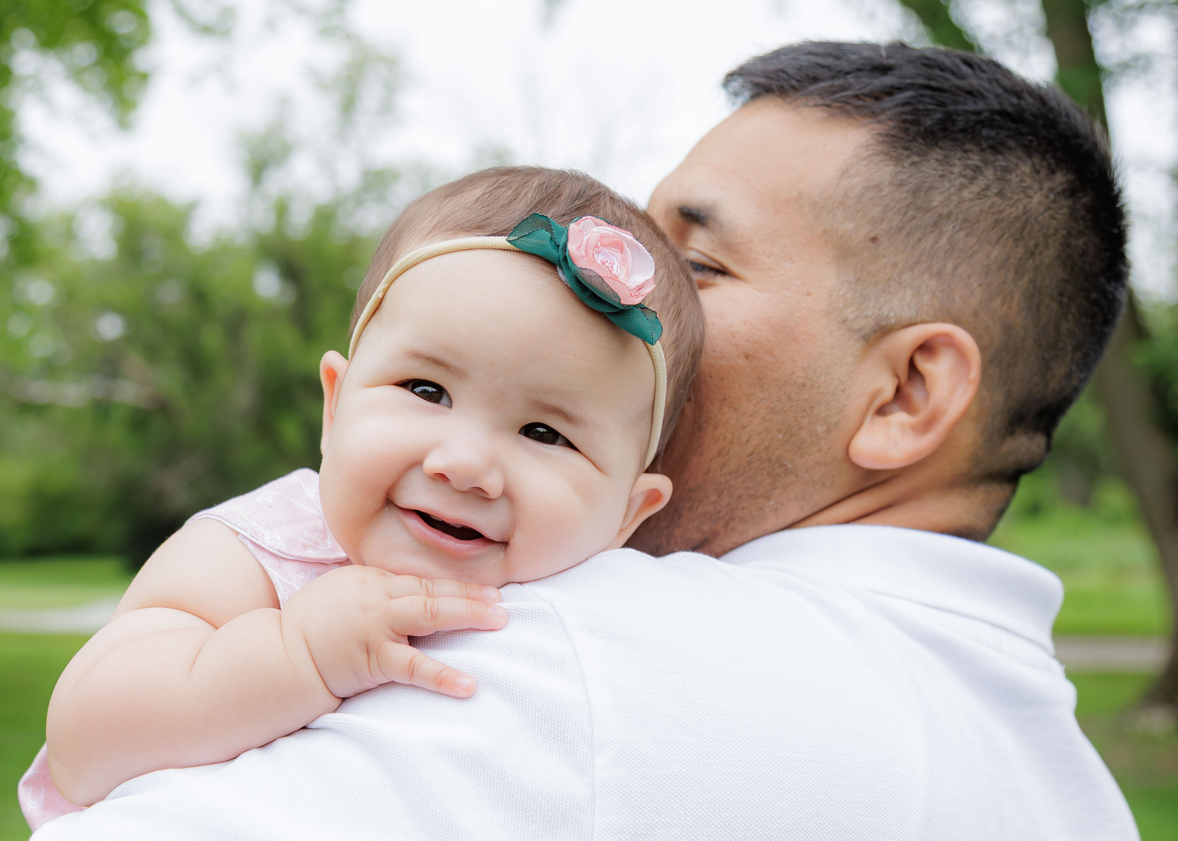 Baby girl resting on dad's shoulder beaming at camera with pink rose headband — IDTPhoto outdoor milestone session Chicagoland