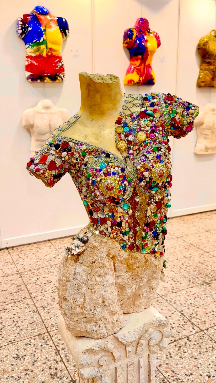Jewel-encrusted bust on stone pedestal with colourful busts on wall behind in gallery
