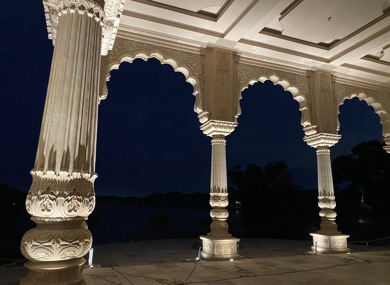Surya Estates colonnade at night — ornate carved Rajasthani columns lit from below by LED Artistry uplights against White Trout Lake
