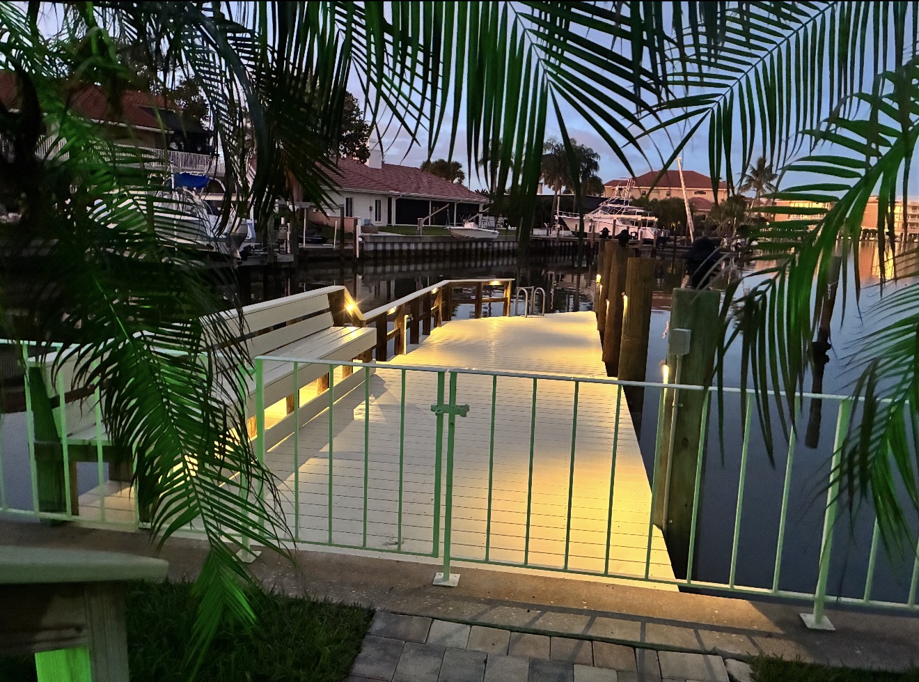 Private waterfront dock with warm LED path and rail lighting at dusk, framed by tropical palms