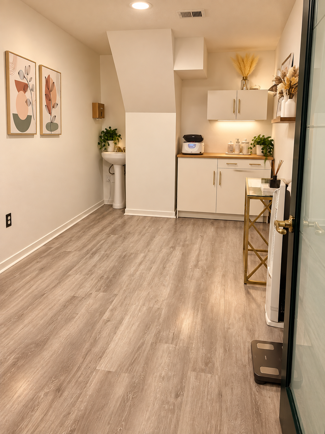 Warm modern spa treatment room with white cabinetry, pedestal sink, gold accents, botanical wall art, and light wood flooring at Posh Salon Suites