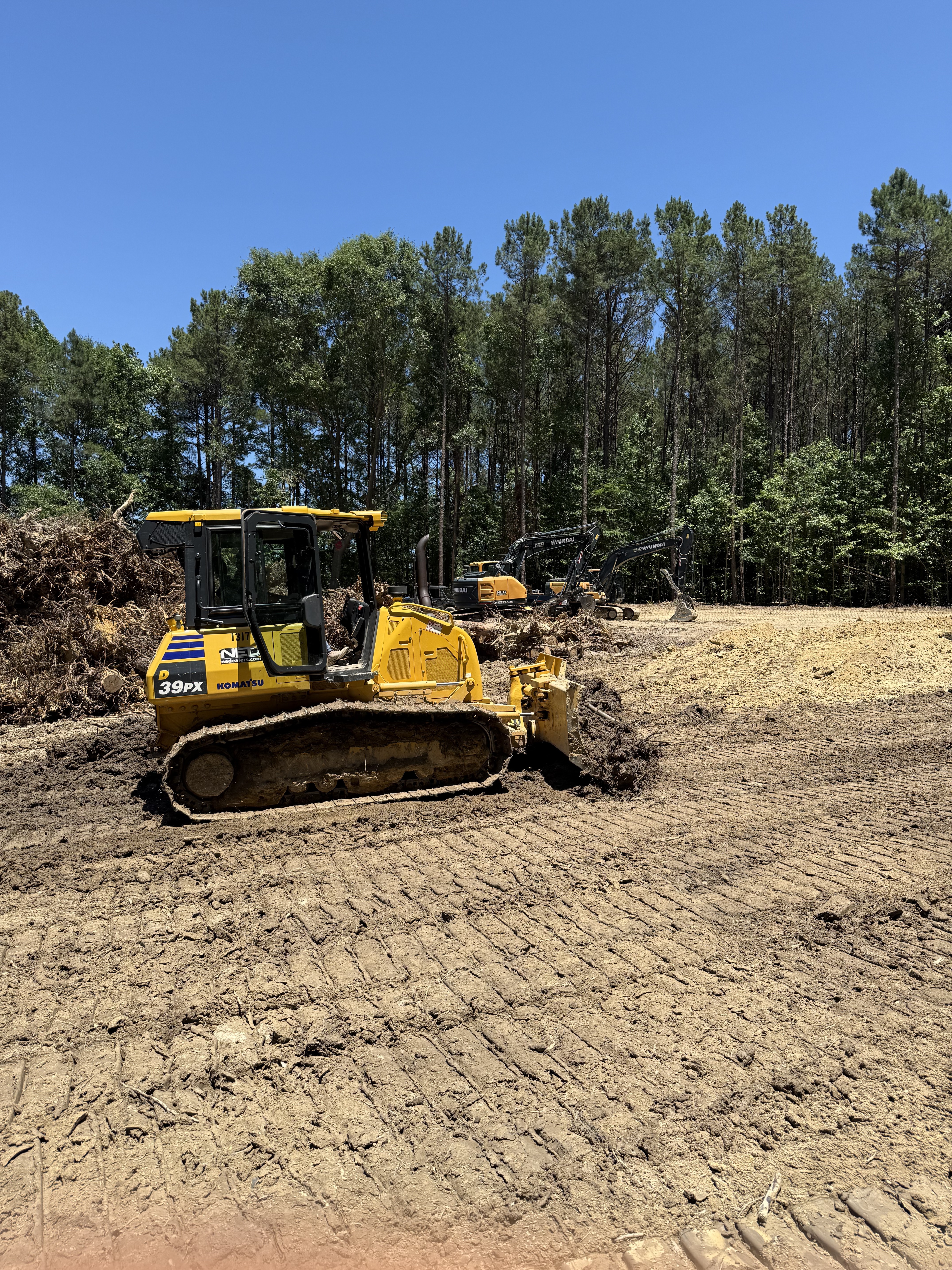 Komatsu D39PX dozer with excavators lined up on a freshly cleared lot with debris piles and pine trees in background, by Stukes Construction