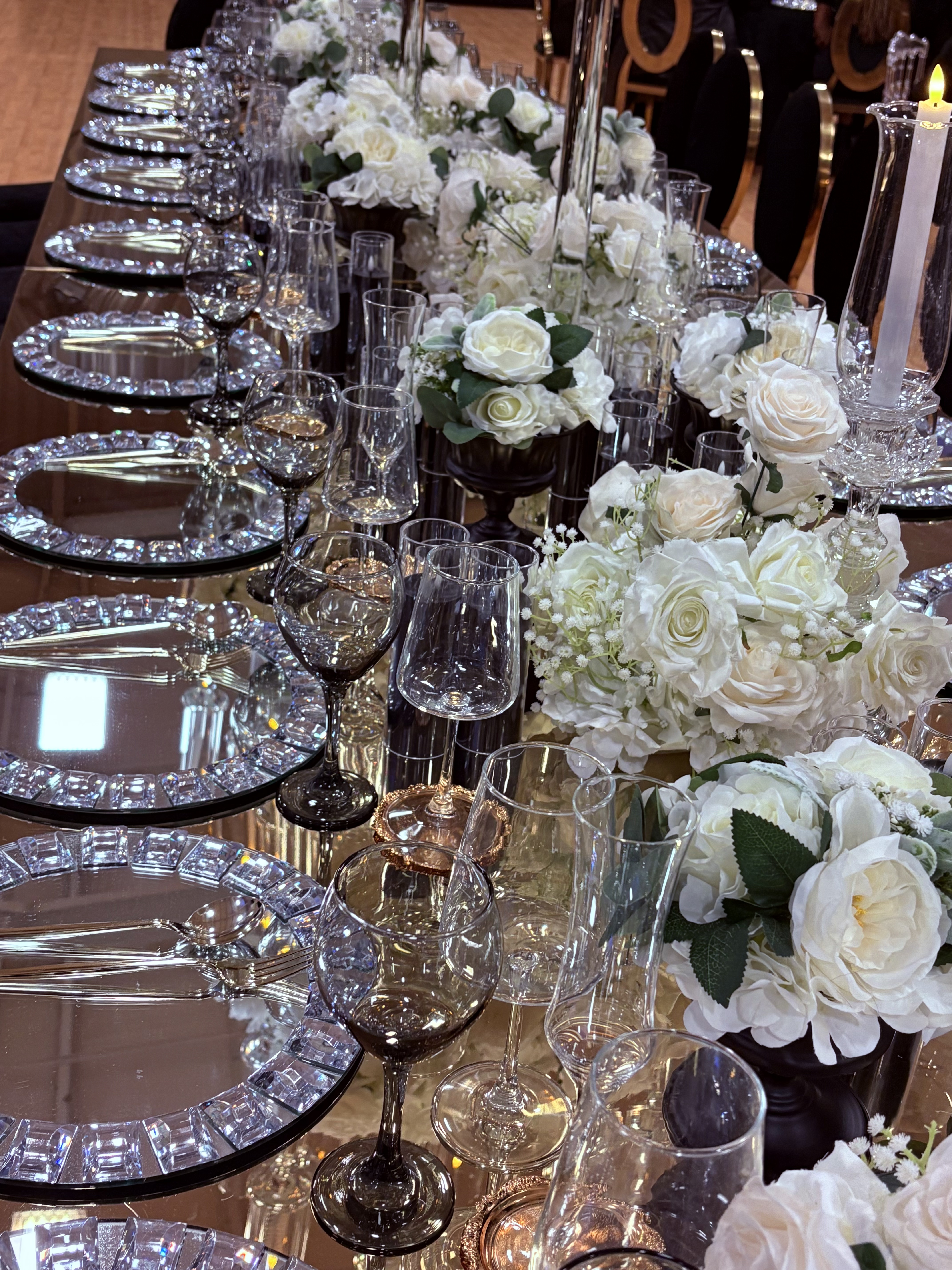 Close-up of mirror table with crystal charger plates, smoky glassware, and white rose floral arrangements