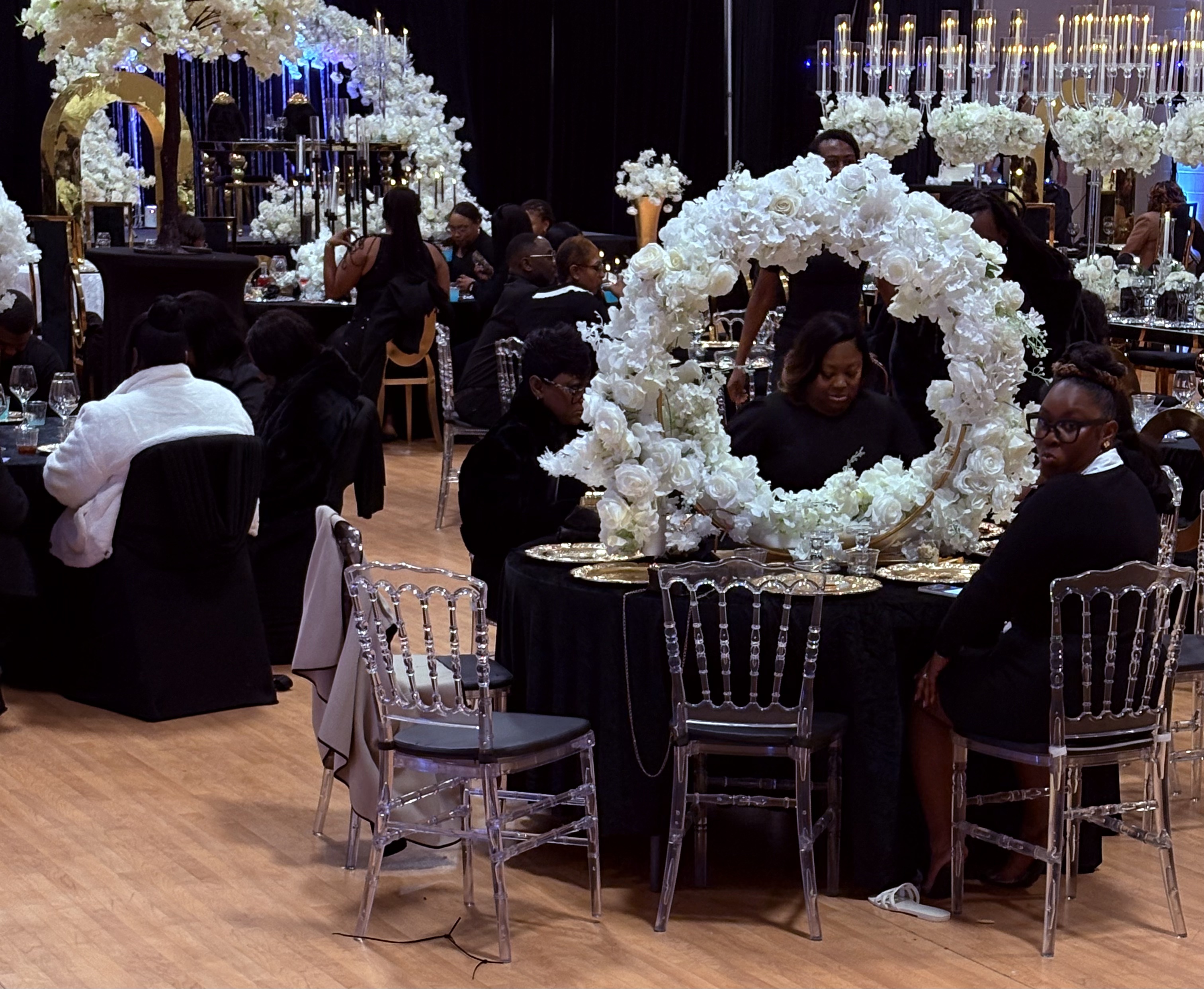 Elegant black and white event with crystal clear chairs, white floral wreath centerpiece, and gold charger plates