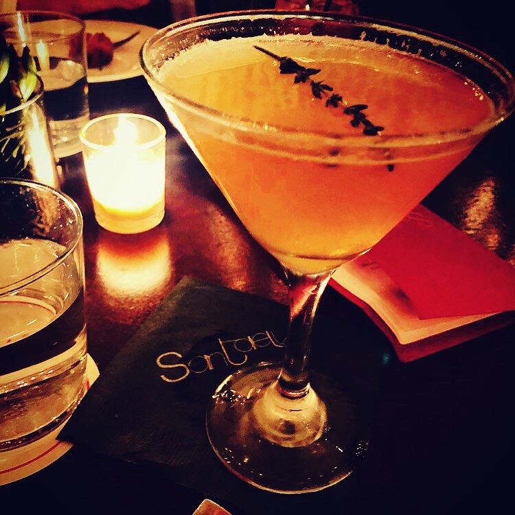 Santaella cocktail — golden martini with lavender garnish on a Santaella-branded napkin, candlelight, warm speakeasy atmosphere
