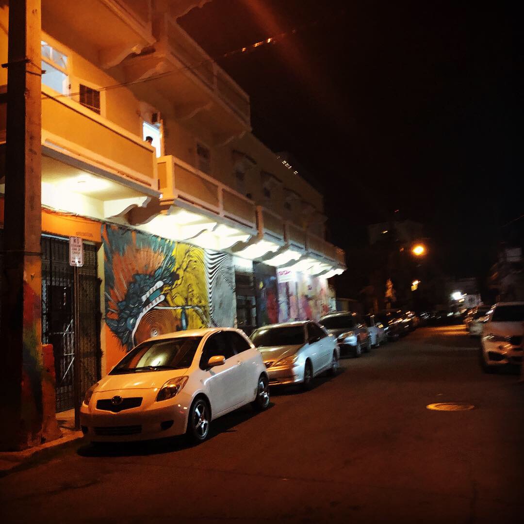 Santurce street art at night — vibrant colorful bird mural on building facade, parked cars, warm street lights, La Cerra area, San Juan, Puerto Rico