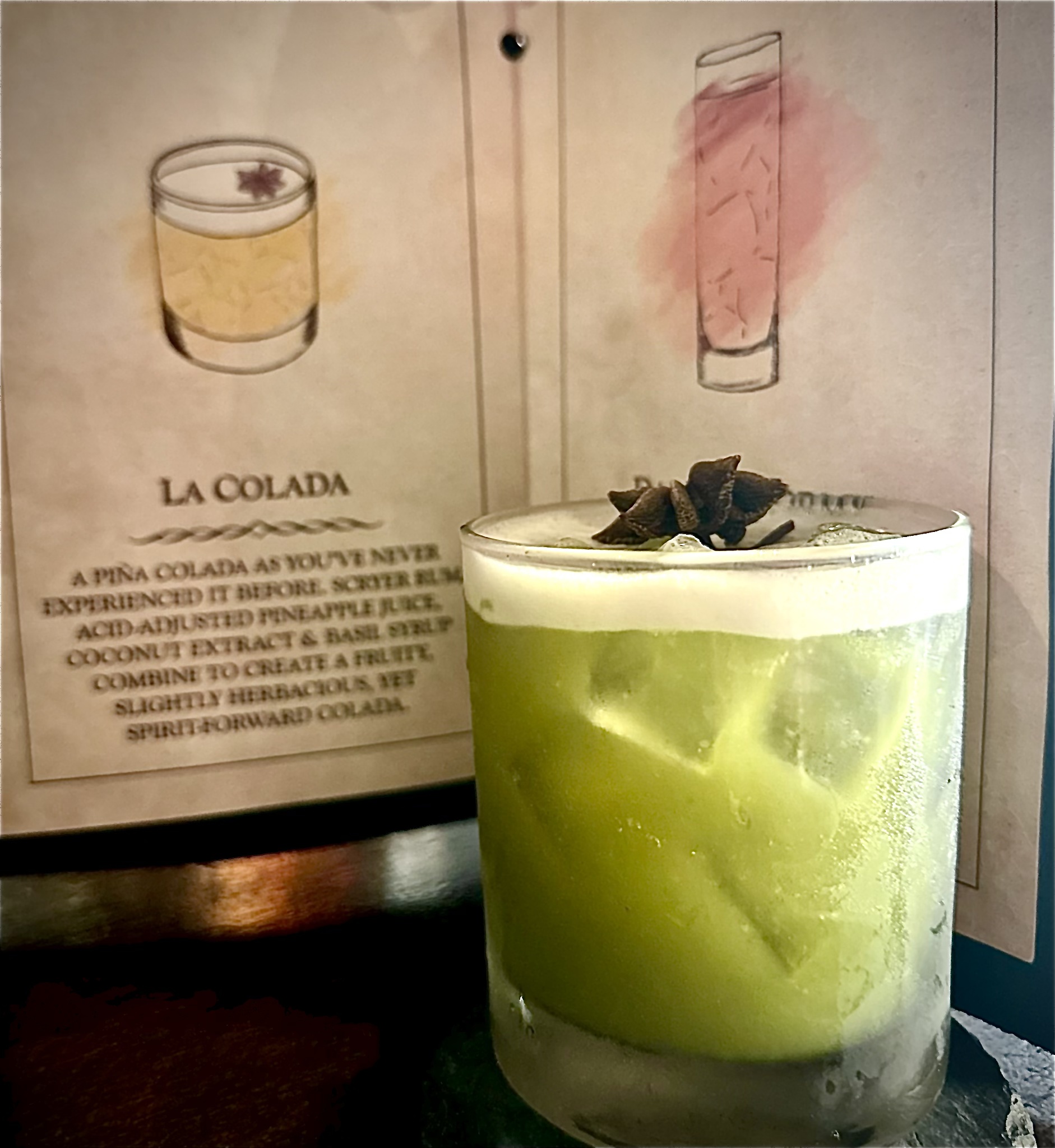 La Colada cocktail at Scryer Rum Barrelhouse \u2014 vibrant green cocktail with star anise garnish in a rocks glass, La Colada menu card in background describing Scryer Rum, acid-adjusted pineapple juice, coconut extract and basil syrup