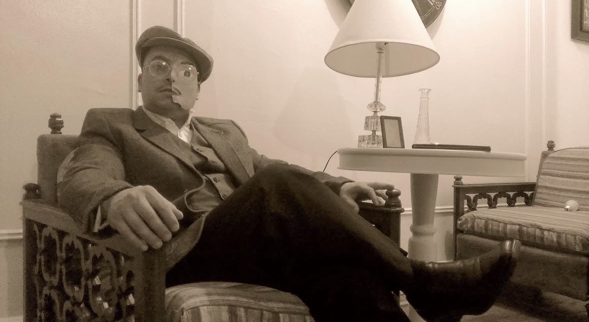 Throwback Halloween β man in 1920s suit, flat cap, and round glasses lounging in an armchair with a cigar β sepia-toned Prohibition-era portrait