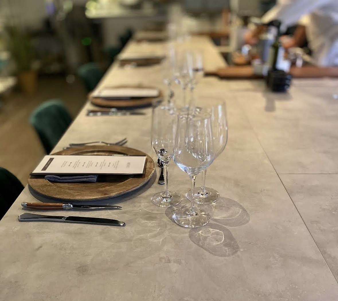 Epicuro table setting — wooden charger plates on concrete table, crystal wine glasses, linen napkins, Laguiole steak knives, menu card