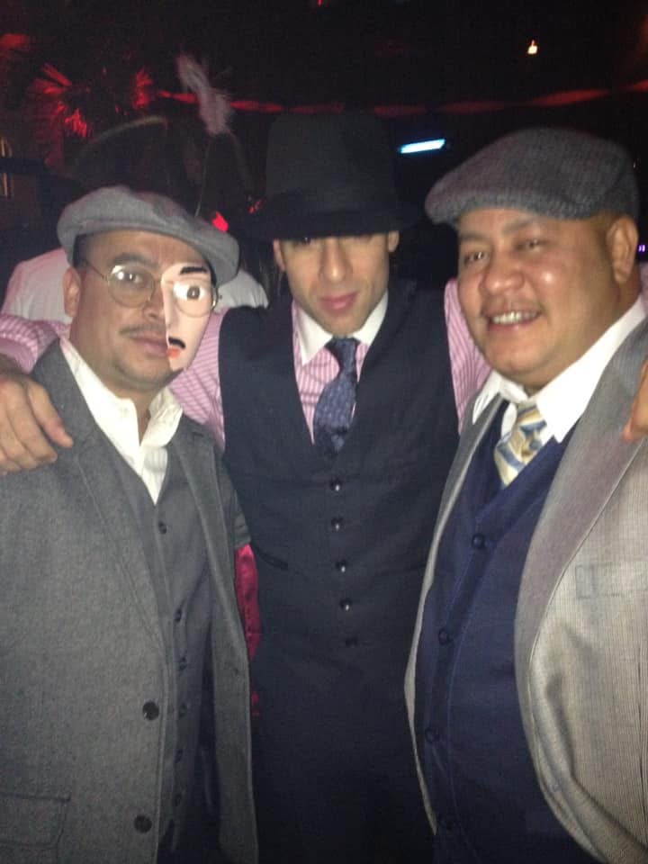 Three men in full 1920s Prohibition attire β flat caps, vests, round glasses β posing together at a Halloween speakeasy party