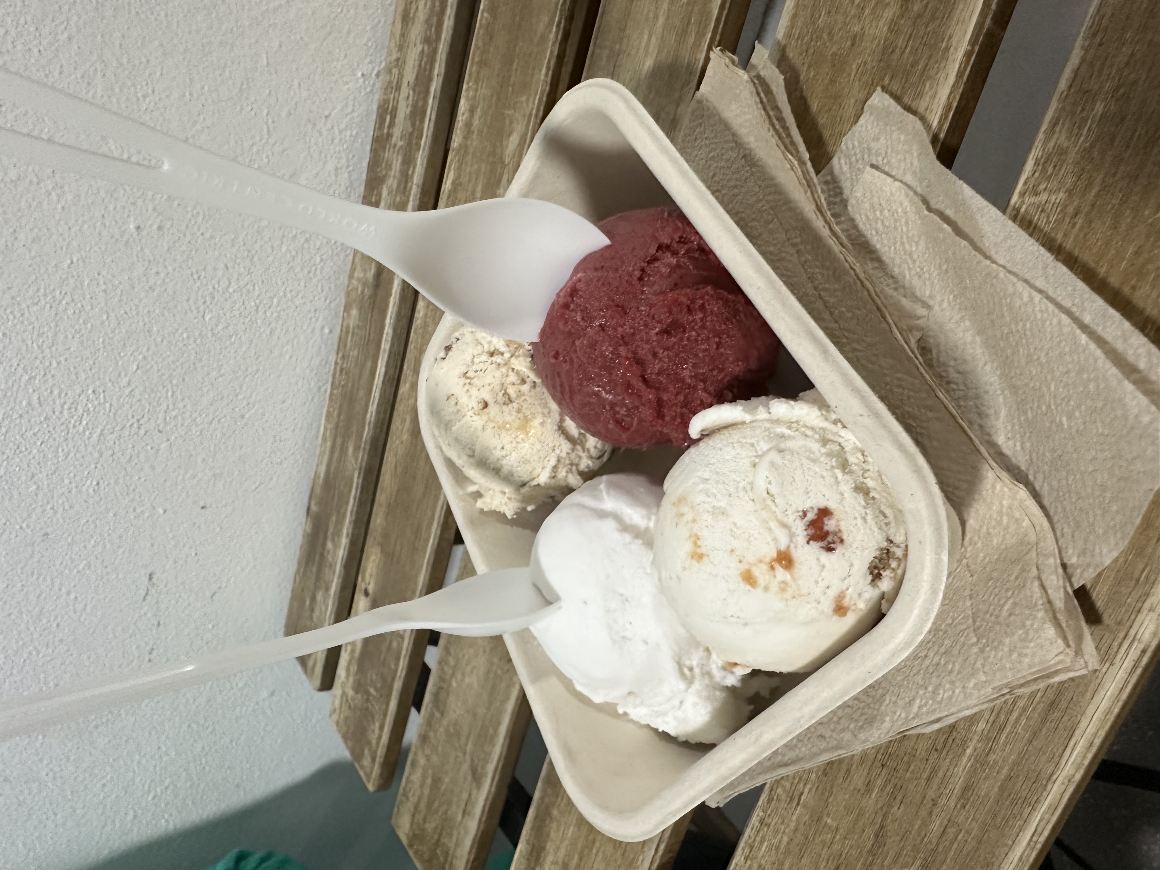 Via Lactea ice cream — four scoops in a compostable container: white coconut, guayaba panetela (deep berry), vanilla, and cream — two spoons, on a wooden slat bench in Santurce, San Juan