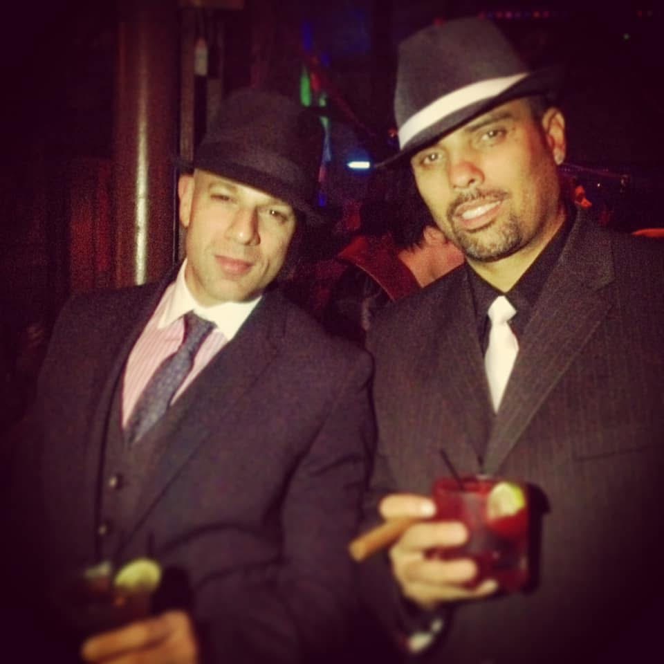 Two men in 1920s Prohibition attire β pinstripe suits, fedora hats, cocktails and cigar β Halloween speakeasy party throwback