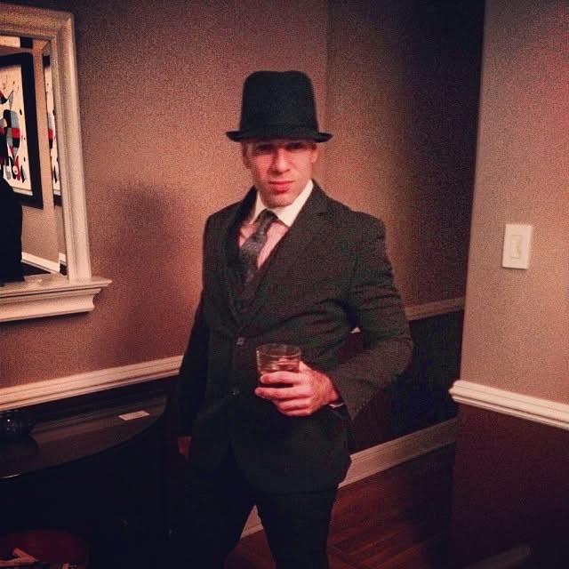Man in dark green three-piece suit, fedora hat, and tie holding a cocktail β Prohibition-era Halloween party throwback