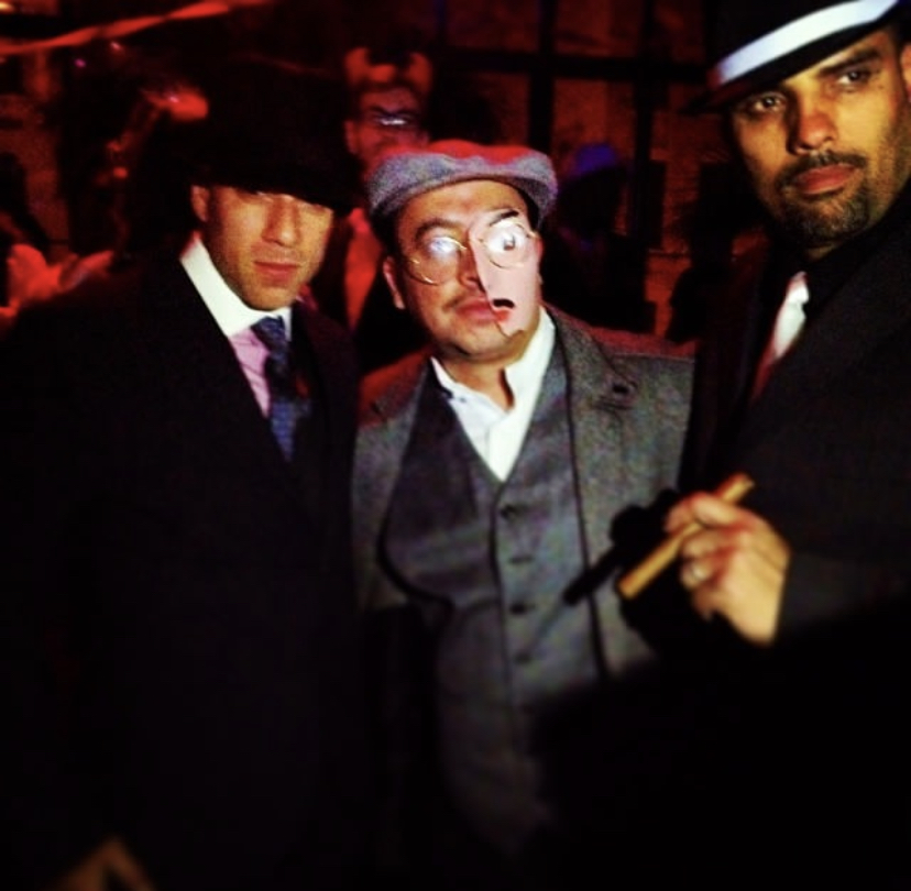 Lucky, Charles, and the crew in full 1920s Prohibition attire β flat caps, vests, round glasses, cigar, dark speakeasy setting β the original Halloween Prohibition party