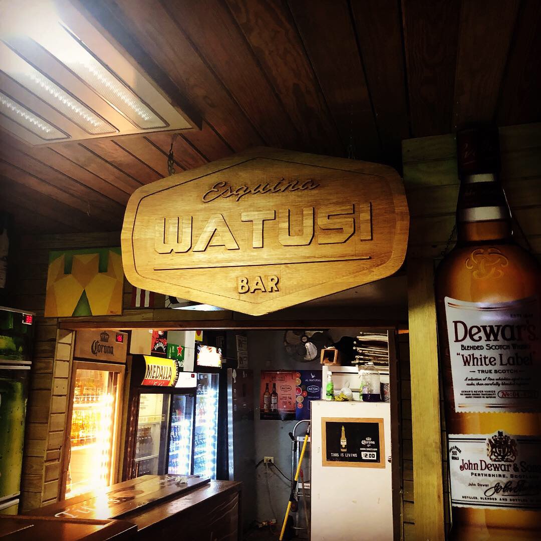 Esquina Watusi Bar — carved wooden sign reading 'Esquina Watusi Bar' hanging from wooden ceiling, Corona and Medalla beer fridges glowing, Dewar's bottle display, classic Puerto Rican neighborhood bar in Santurce