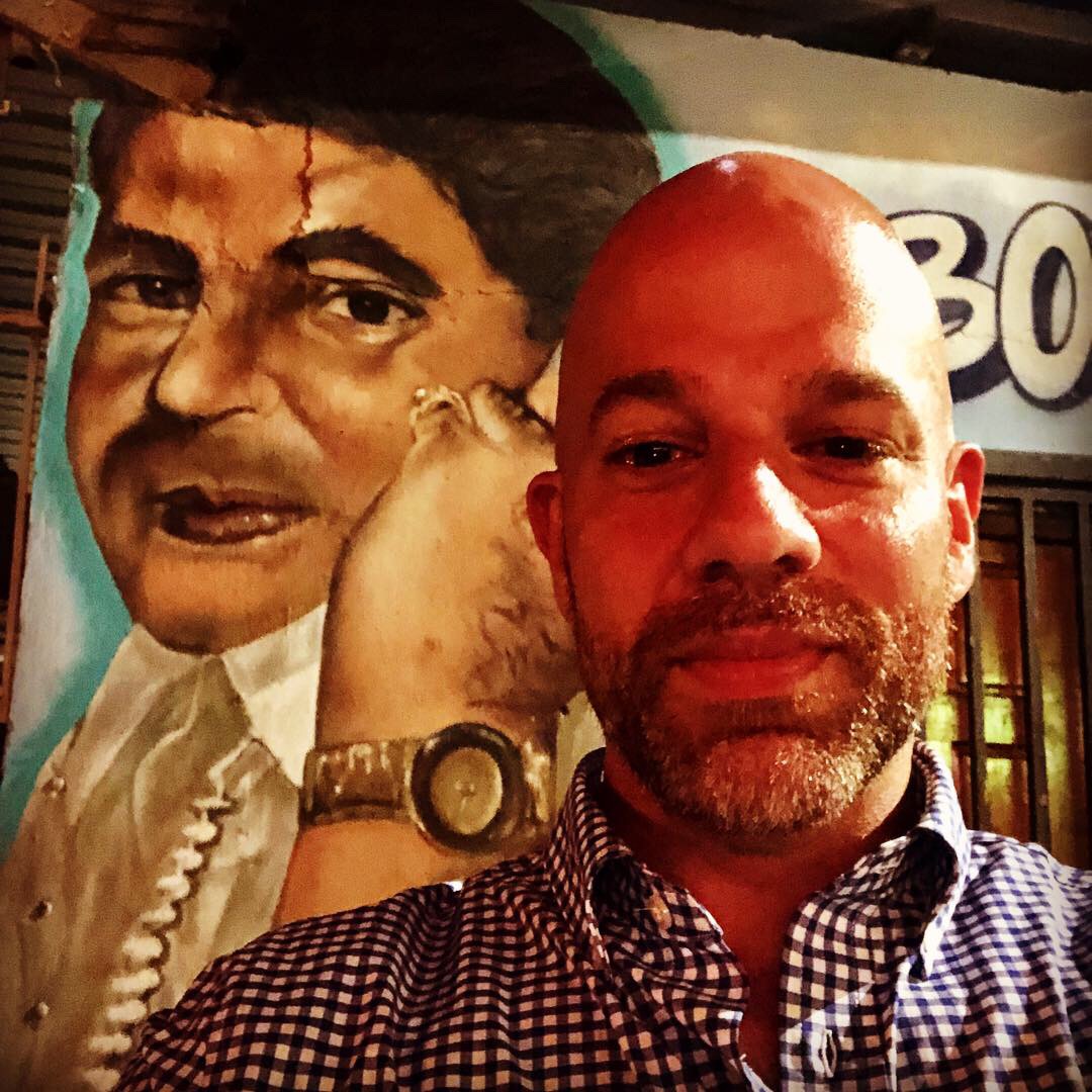 Xavier Marzan selfie in front of a large mural portrait on La Cerra Street in Santurce — wearing a checkered shirt, warm night lighting, Puerto Rican street culture