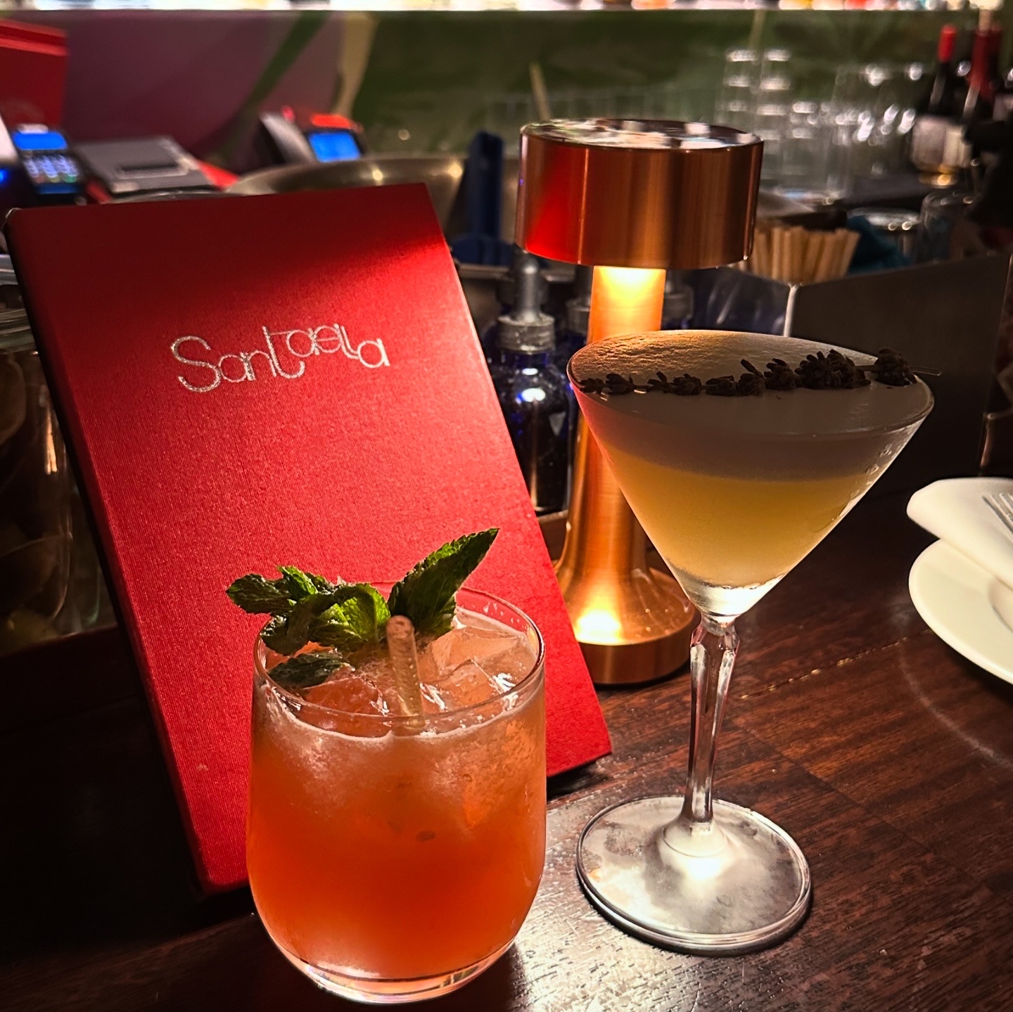 Santaella cocktails — two craft cocktails beside the red Santaella menu, one with mint garnish, one martini with lavender, warm amber bar lighting
