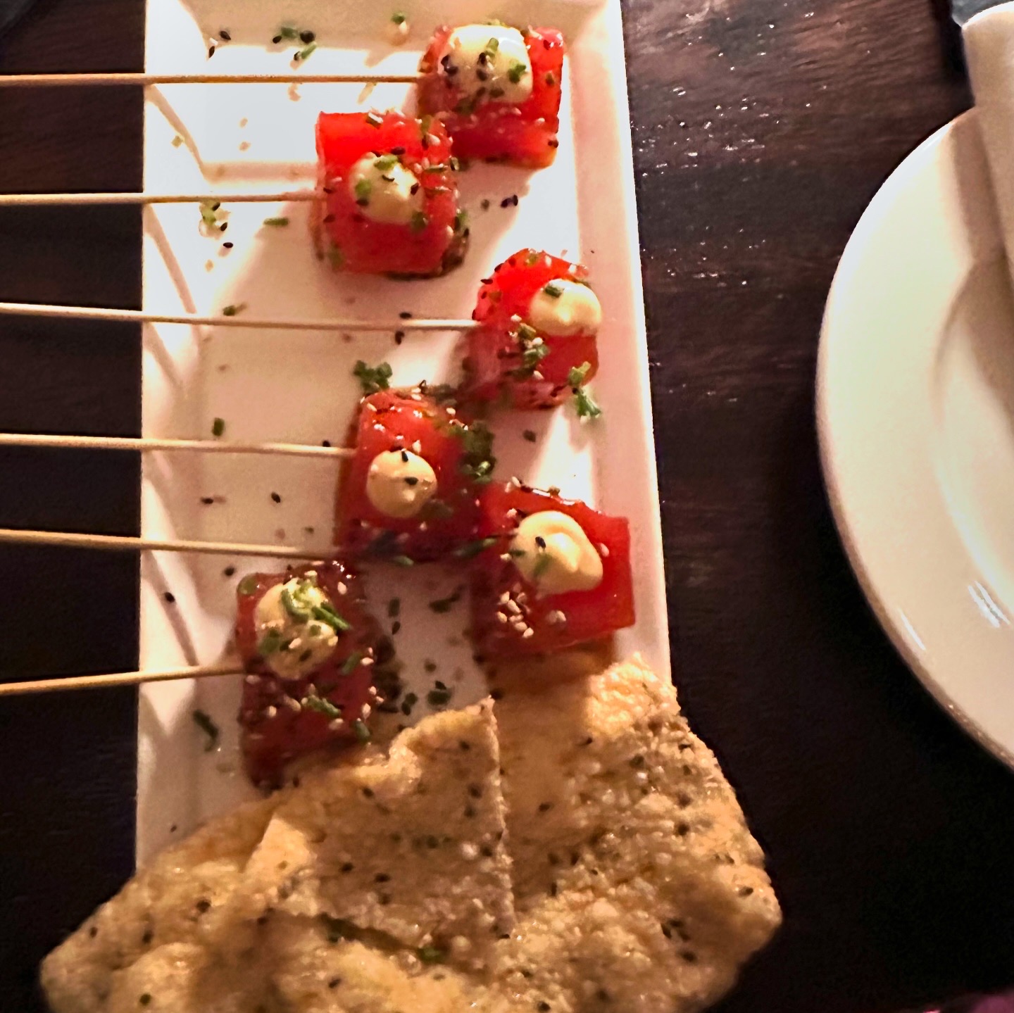 Santaella tuna tartare appetizer — vibrant red tuna cubes with cream dots, sesame seeds, chives, and crispy crackers on a white rectangular plate