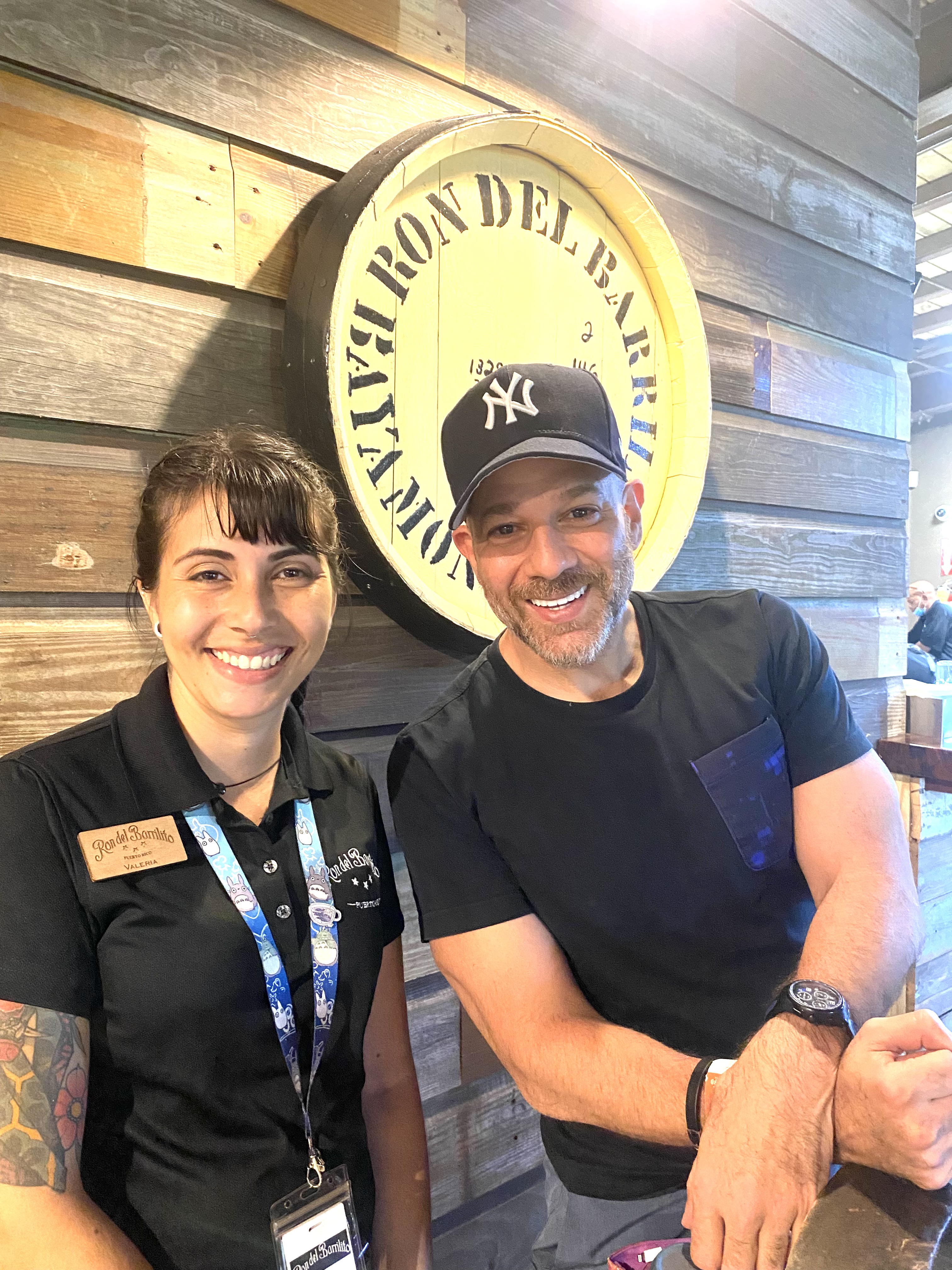 Xavier at Ron del Barrilito rum distillery in Puerto Rico with Valeria — the iconic barrel sign on the wood-paneled wall