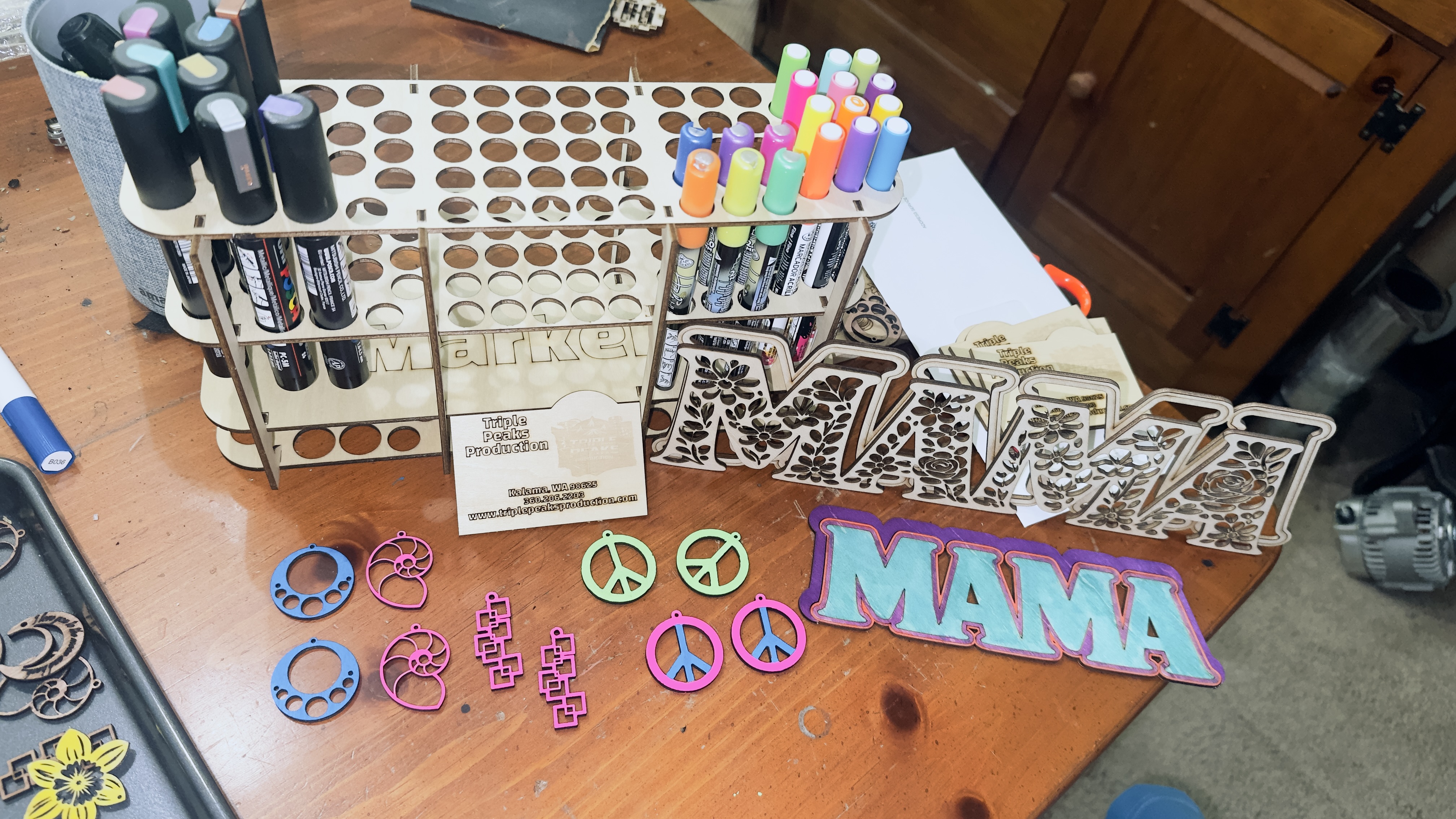 Assorted laser-cut items including colorful acrylic earrings, peace sign pendants, layered MAMA sign, floral letter cutouts, and a marker organizer rack
