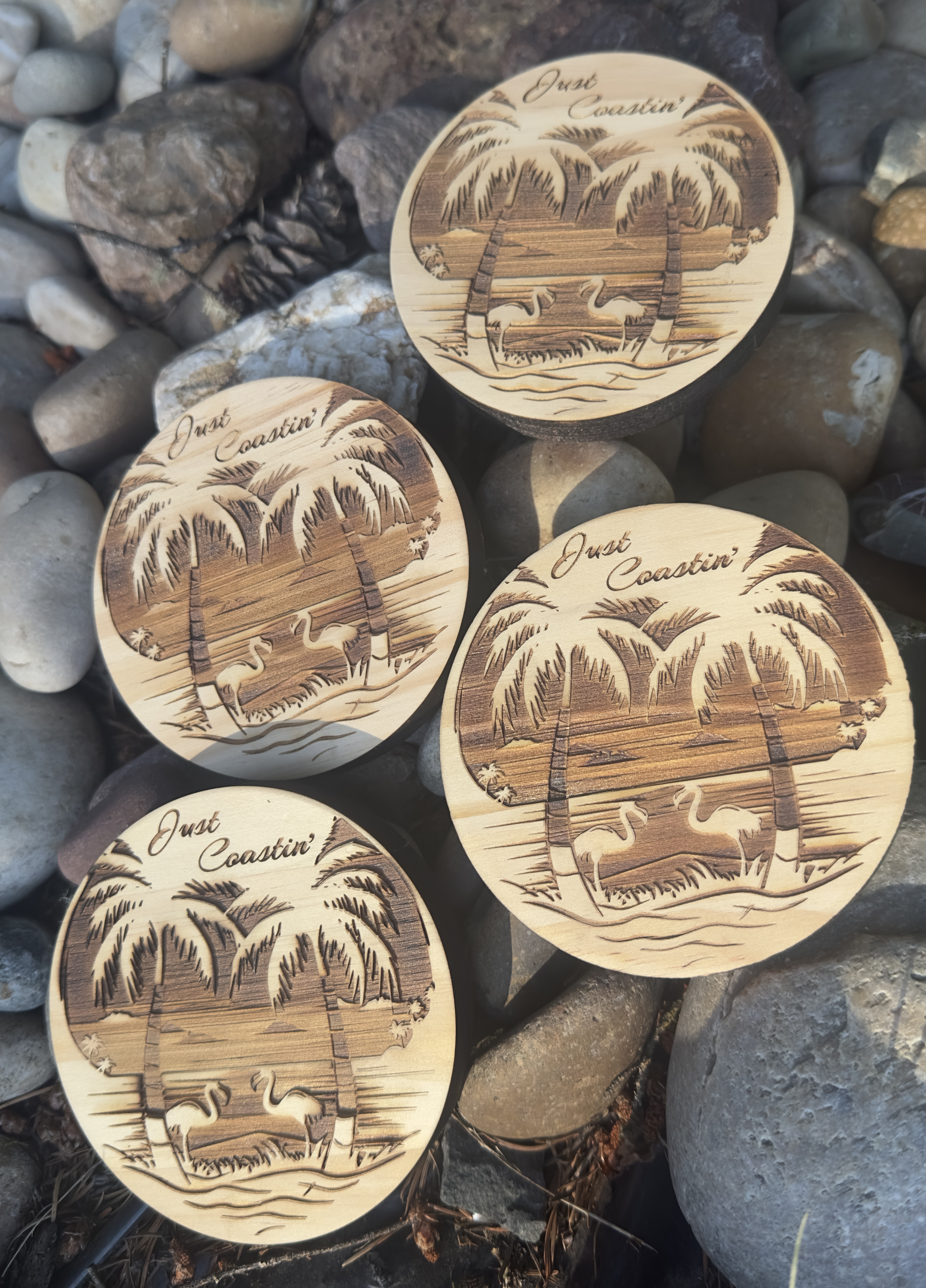 Laser-engraved wooden coasters with palm tree beach scene and 'Just Coastin'' text on a rocky background