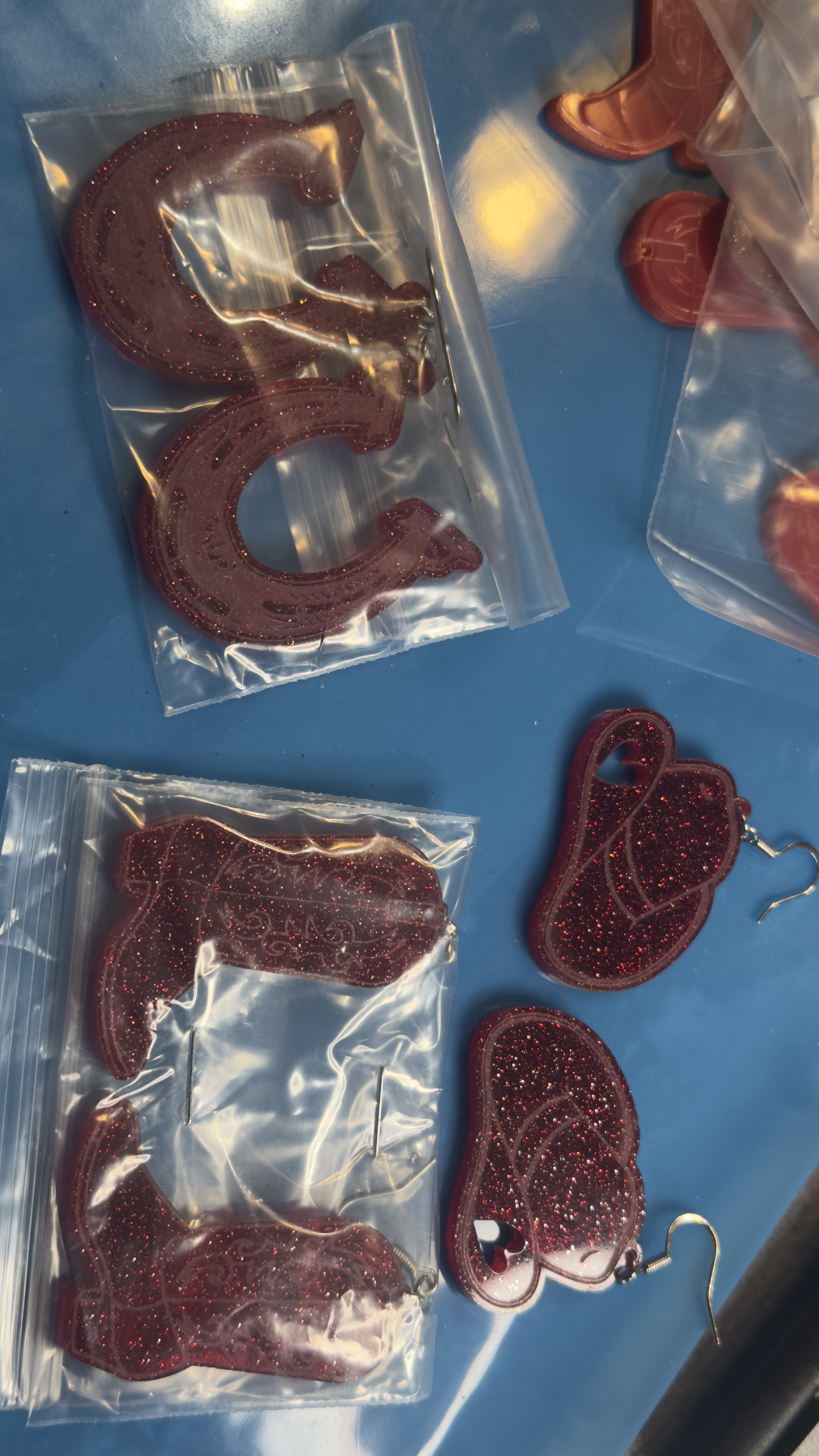 Laser-cut glitter acrylic western earrings and keychains featuring cowboy boots, horseshoes, and cowboy hat designs in deep red sparkle