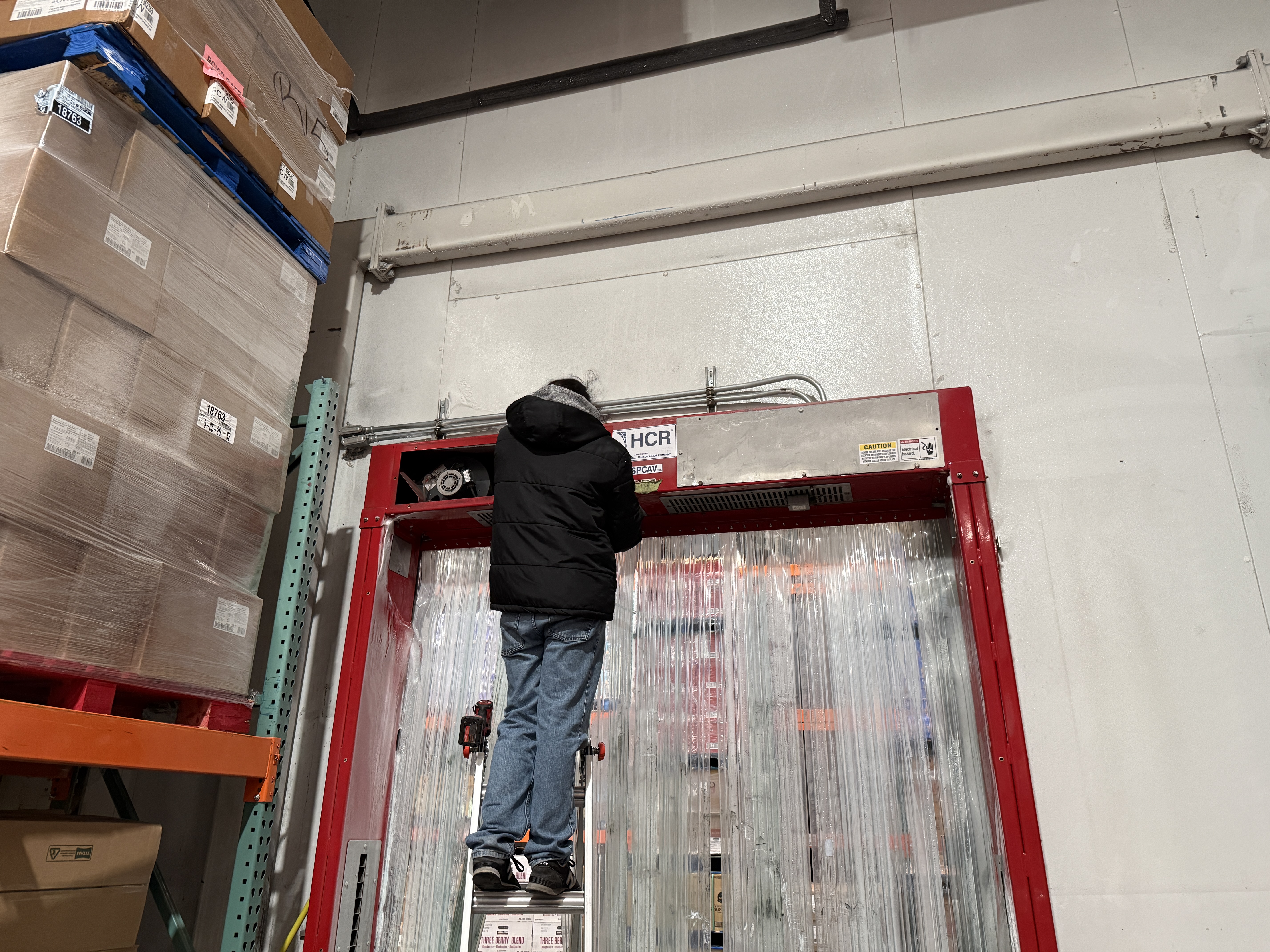 Cal Gaskets technician on ladder installing strip curtains in a warehouse cooler doorway