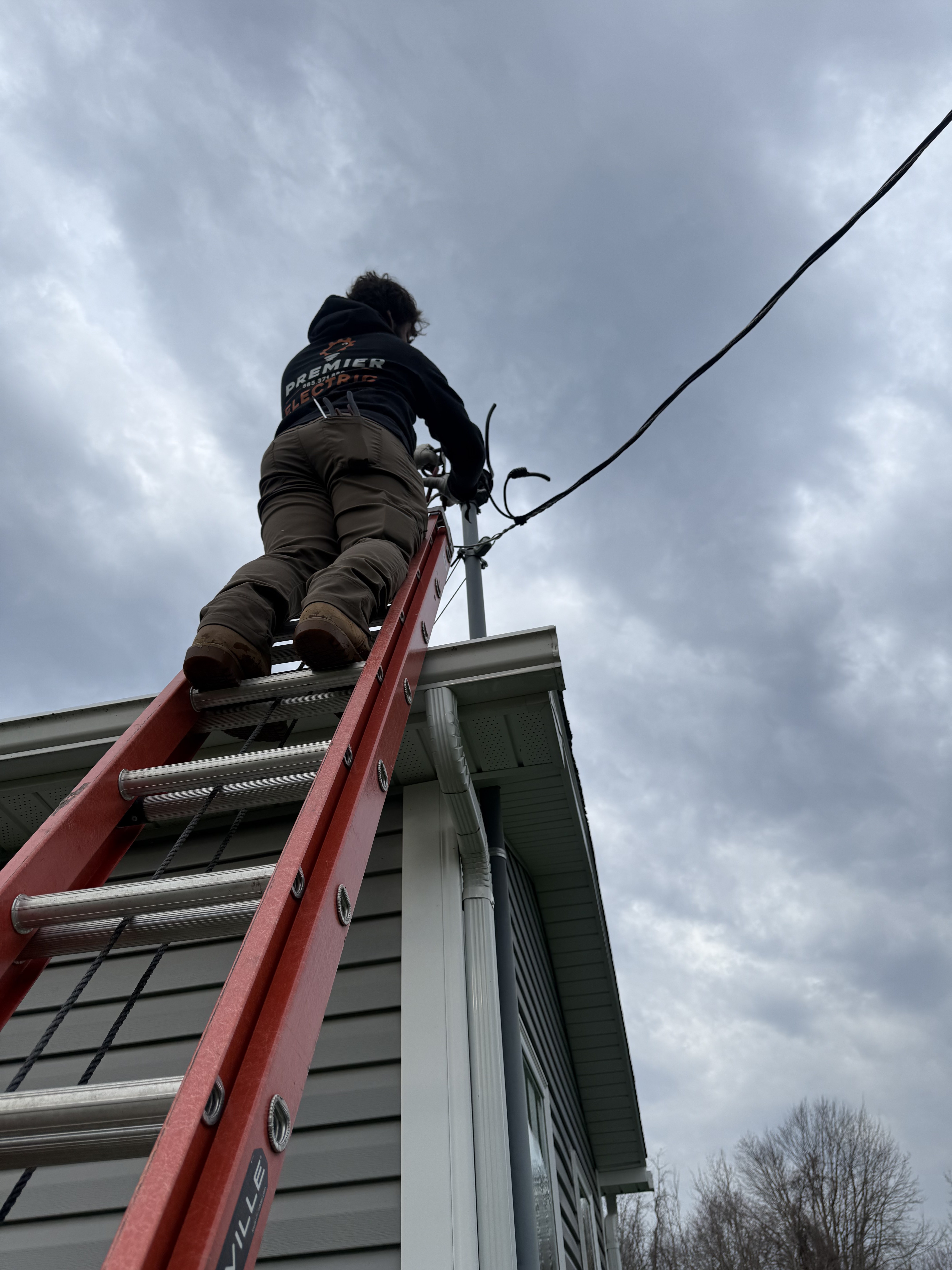 Premier Alarms and Electric technician climbing ladder to work on exterior service entrance at roofline โ Monroe County NY