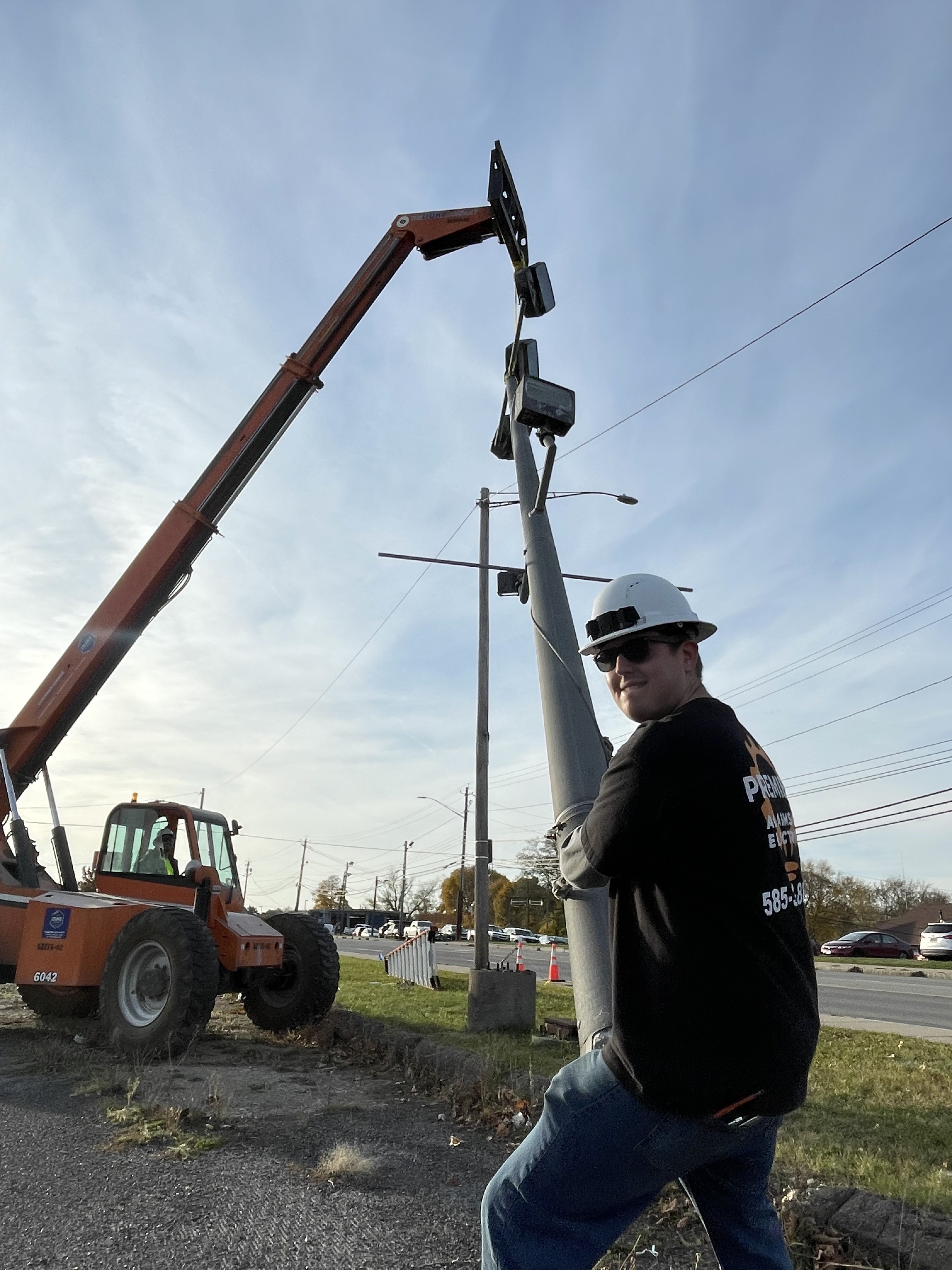Premier Alarms and Electric crew member in hard hat with telehandler installing commercial light pole on roadside โ Monroe County NY